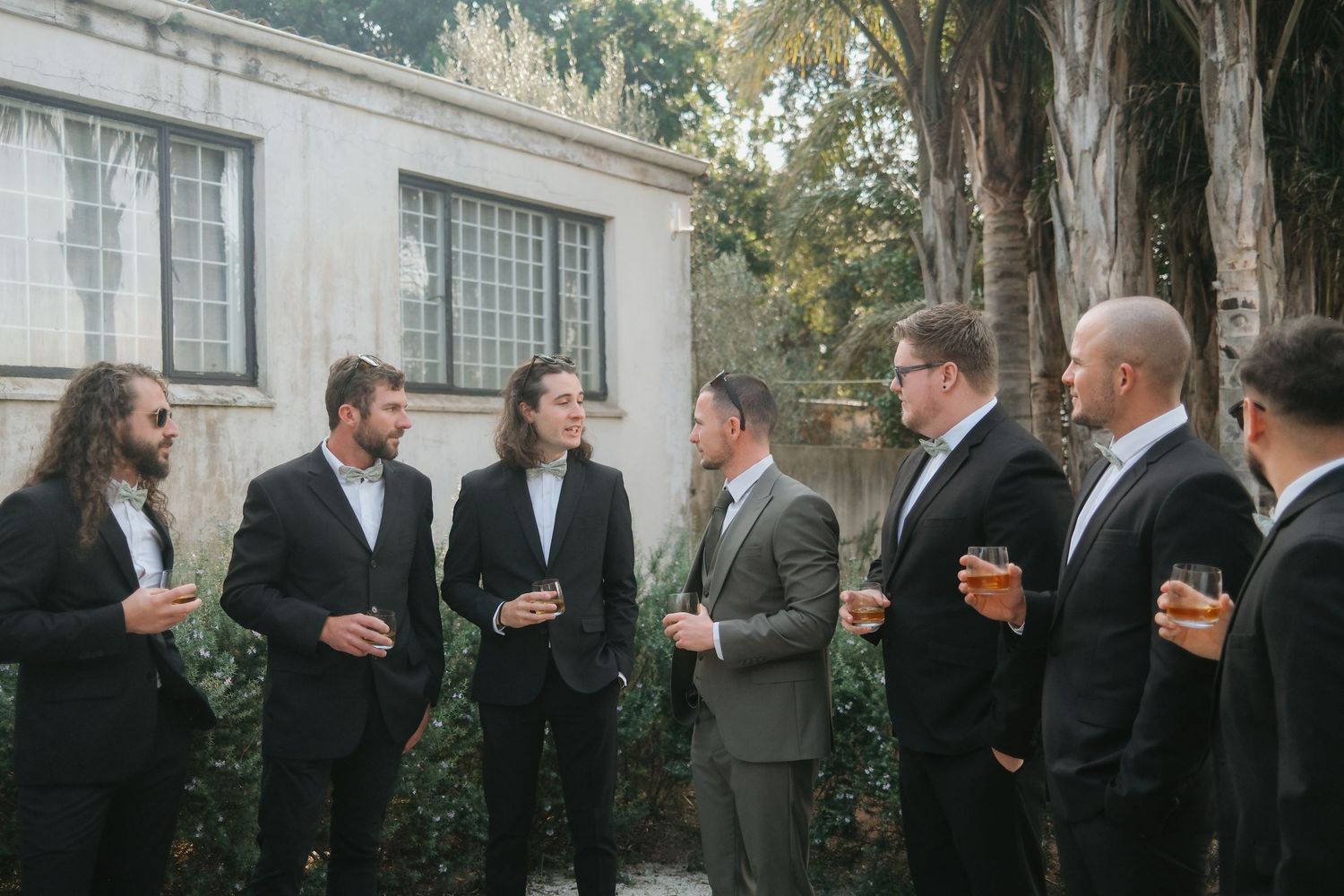 Groomsmen in dark suits gather outdoors conversing, holding drinks near a stone building with lush greenery.