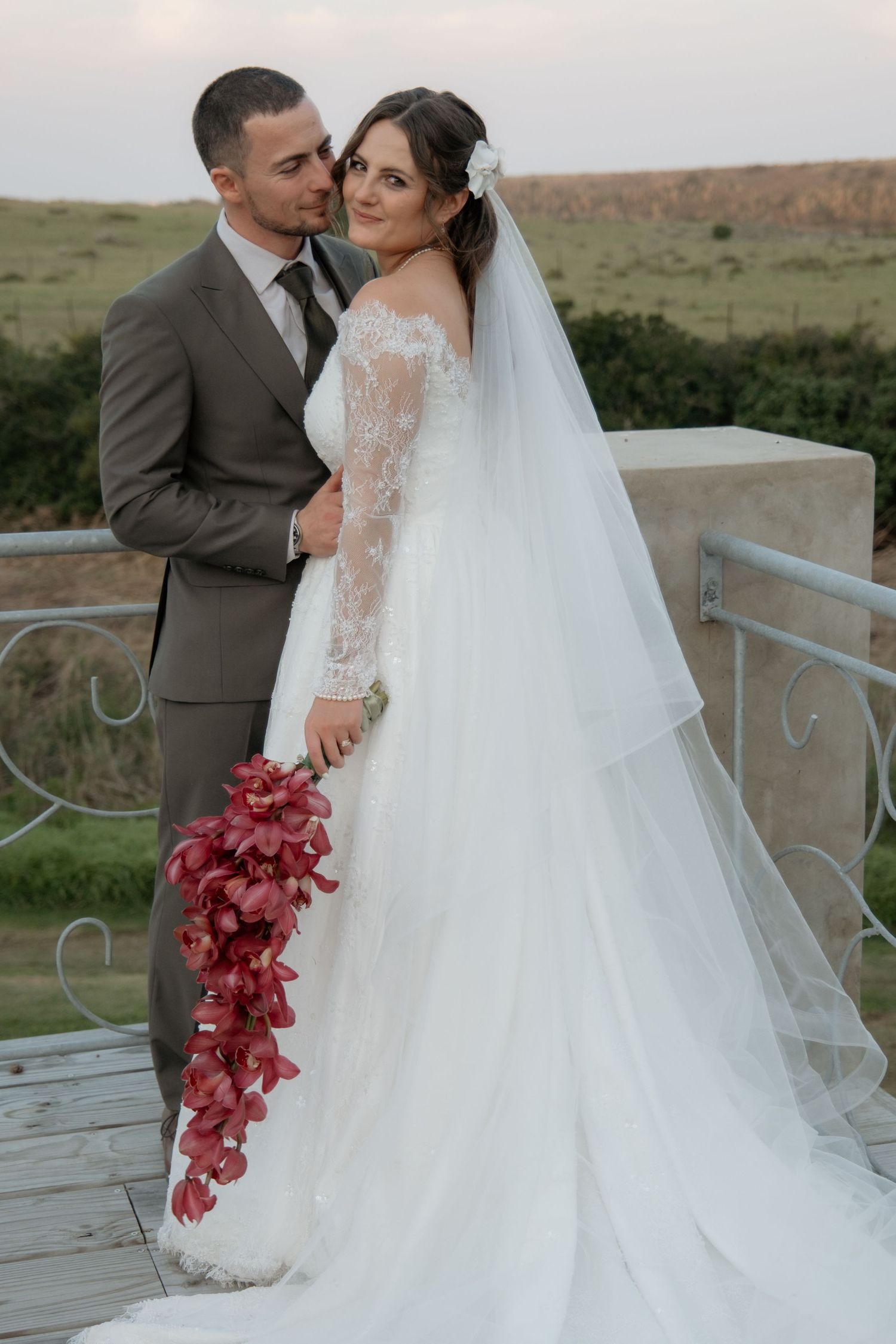 Bride and groom portrait outdoors, bride in lace wedding gown with red cascading bouquet at sunset.