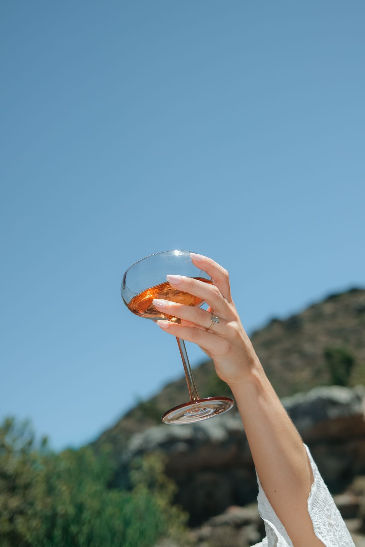 A woman's hand elegantly holds a wine glass with amber liquid against a clear blue sky and mountain backdrop.