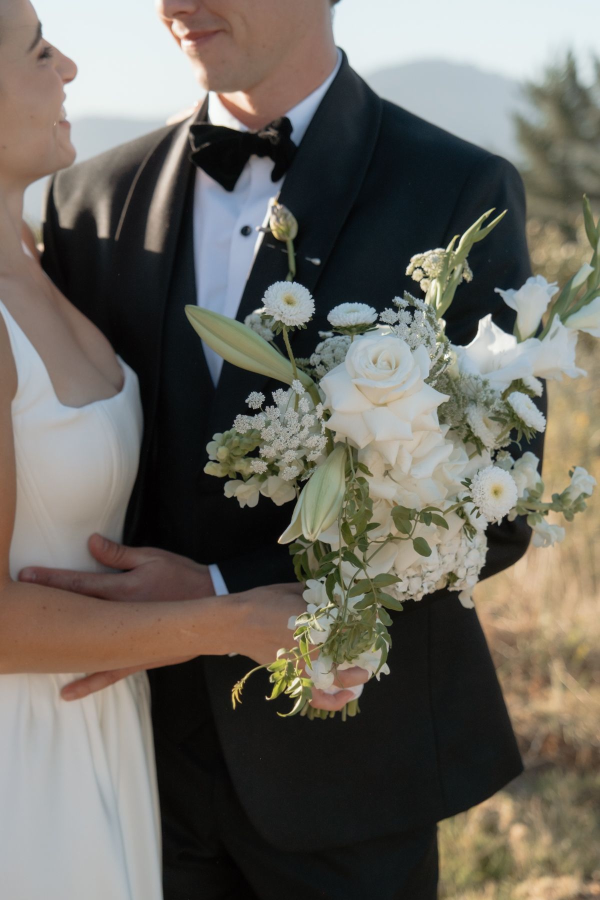 Bride and groom pose together holding elegant white rose and lily wedding bouquet outdoors.