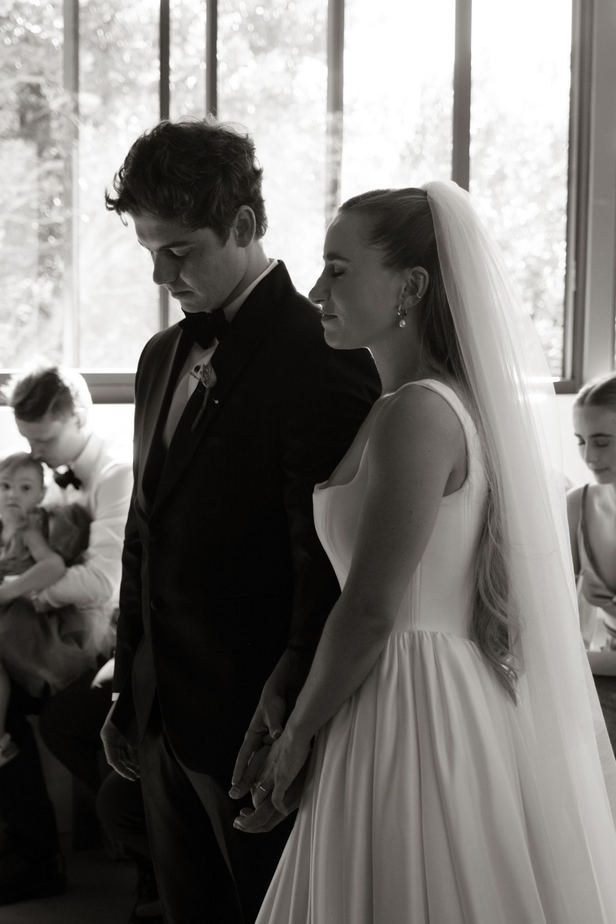 Black and white wedding photo of bride in veil and groom in tuxedo holding hands during ceremony.