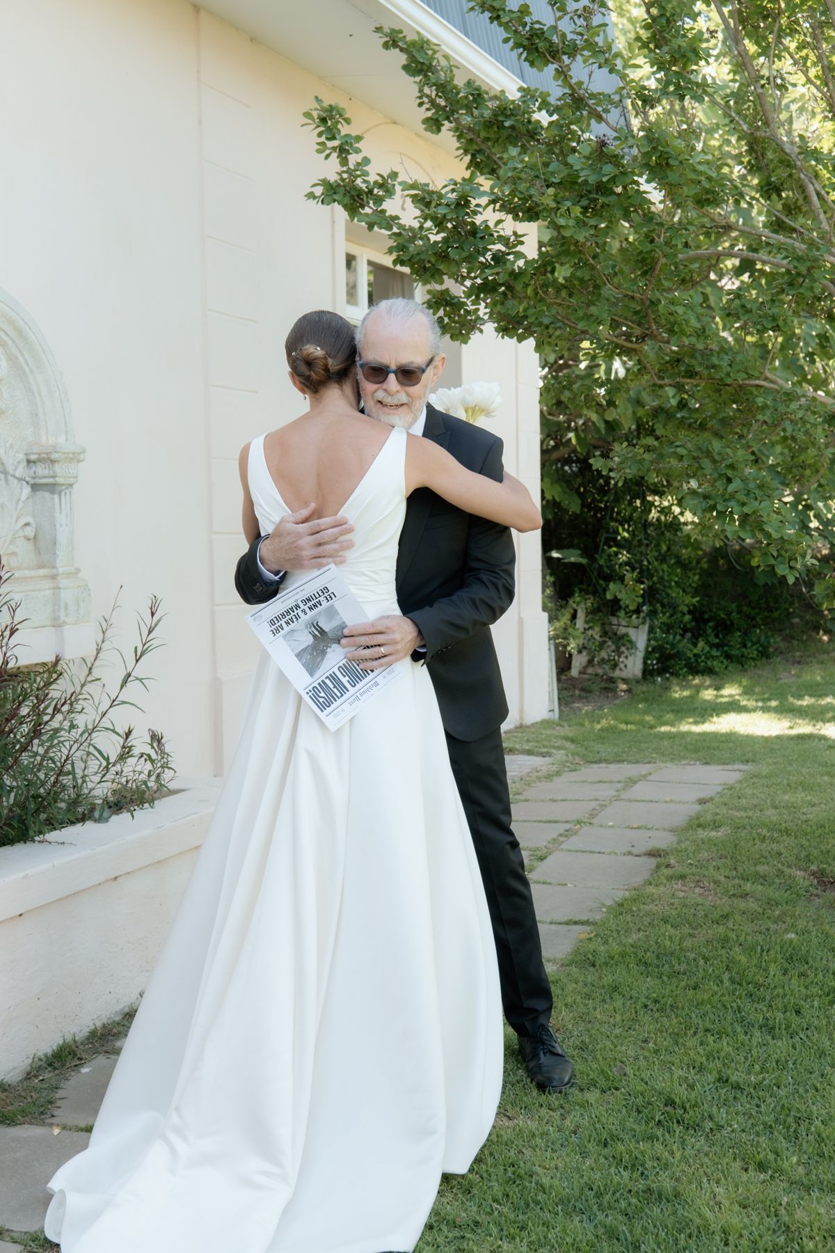 Bride in elegant white gown shares heartfelt hug with older gentleman in black suit outdoors.