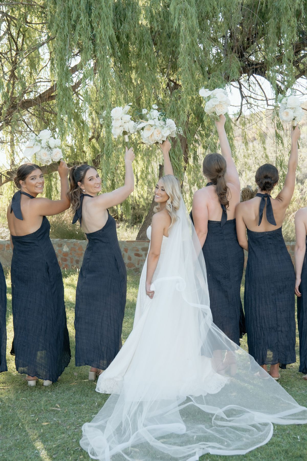Bride in white gown with bridesmaids in navy dresses raising white bouquets under willow tree.