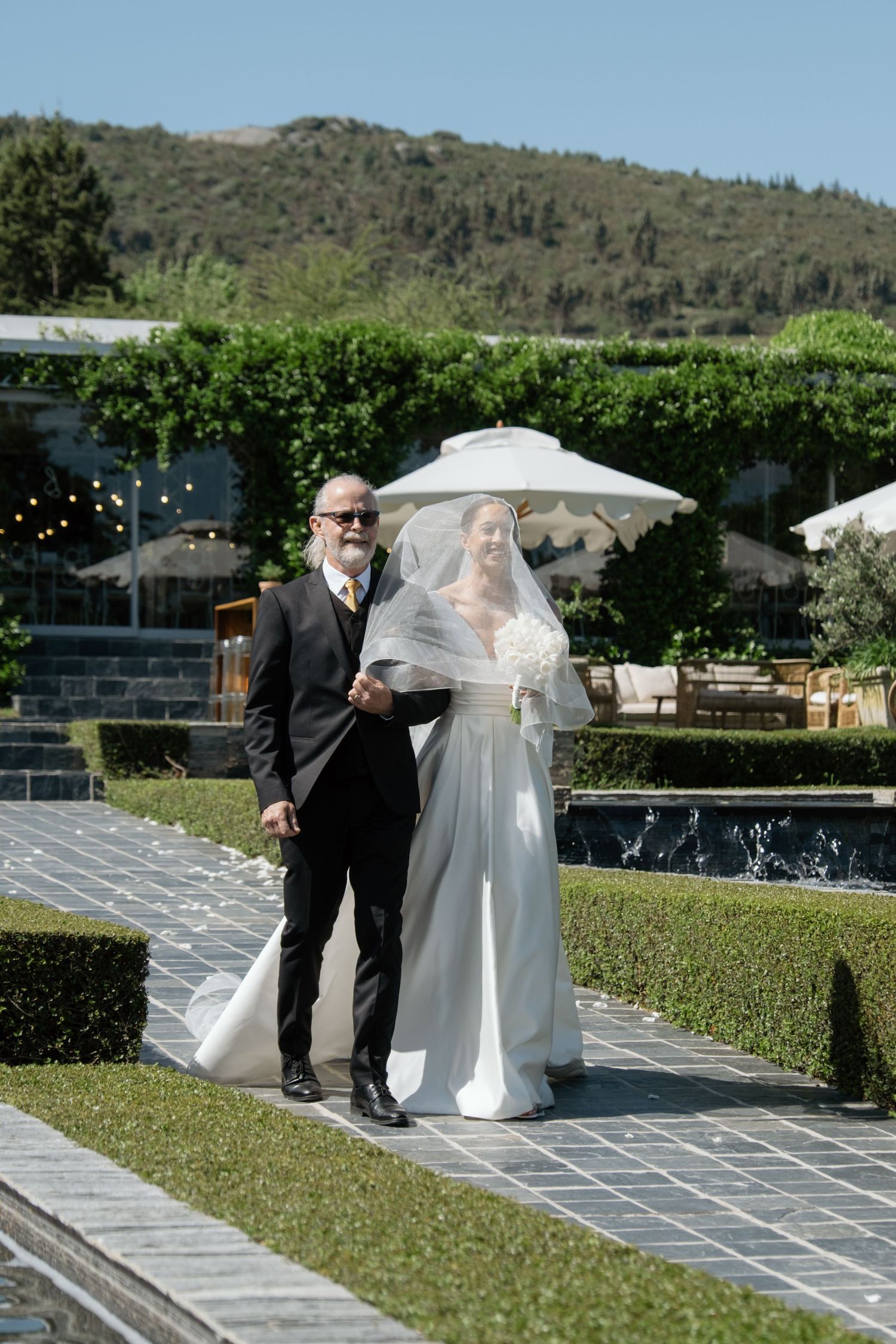 Bride in white gown walks with older man in black suit through elegant garden venue on sunny wedding day.