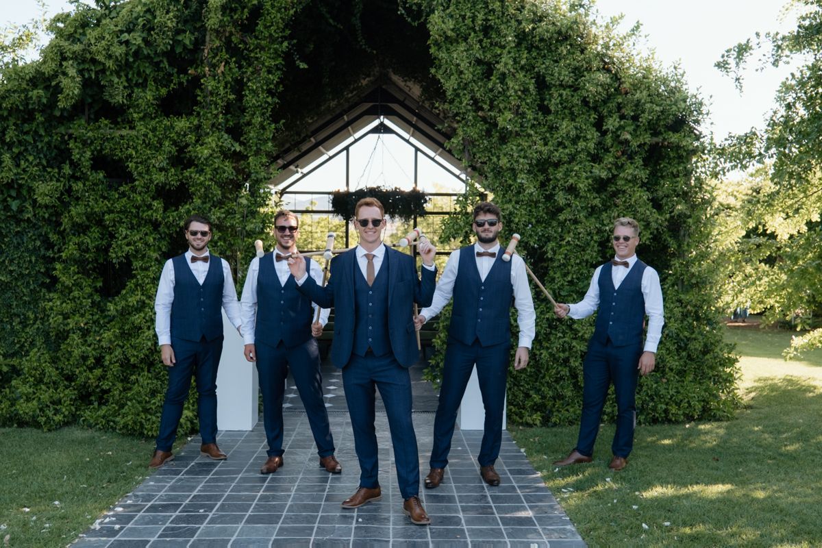 Groom and groomsmen in matching navy suits pose confidently in front of a garden wedding arch.