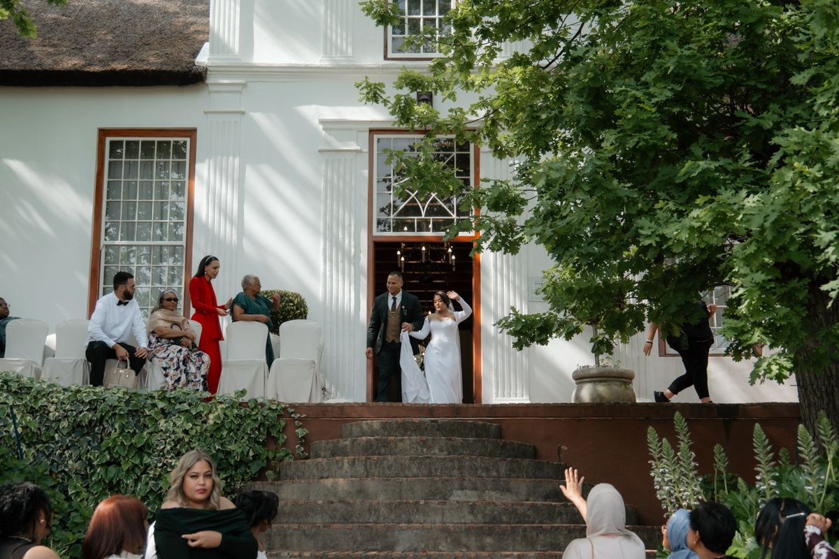 Bride and groom exit historic white building venue as wedding guests celebrate on cobblestone steps outside.