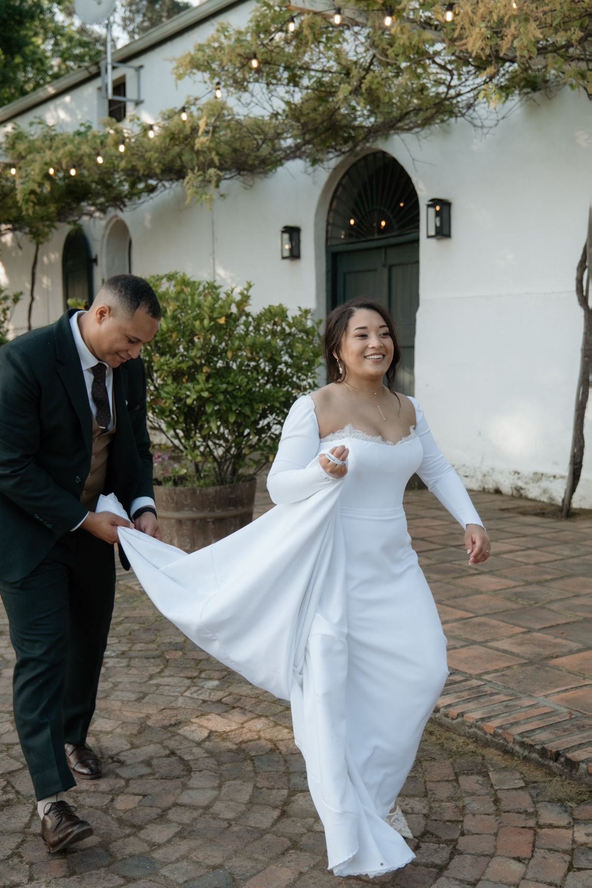 Smiling bride in white long-sleeve gown walks with groom outside white venue adorned with string lights.