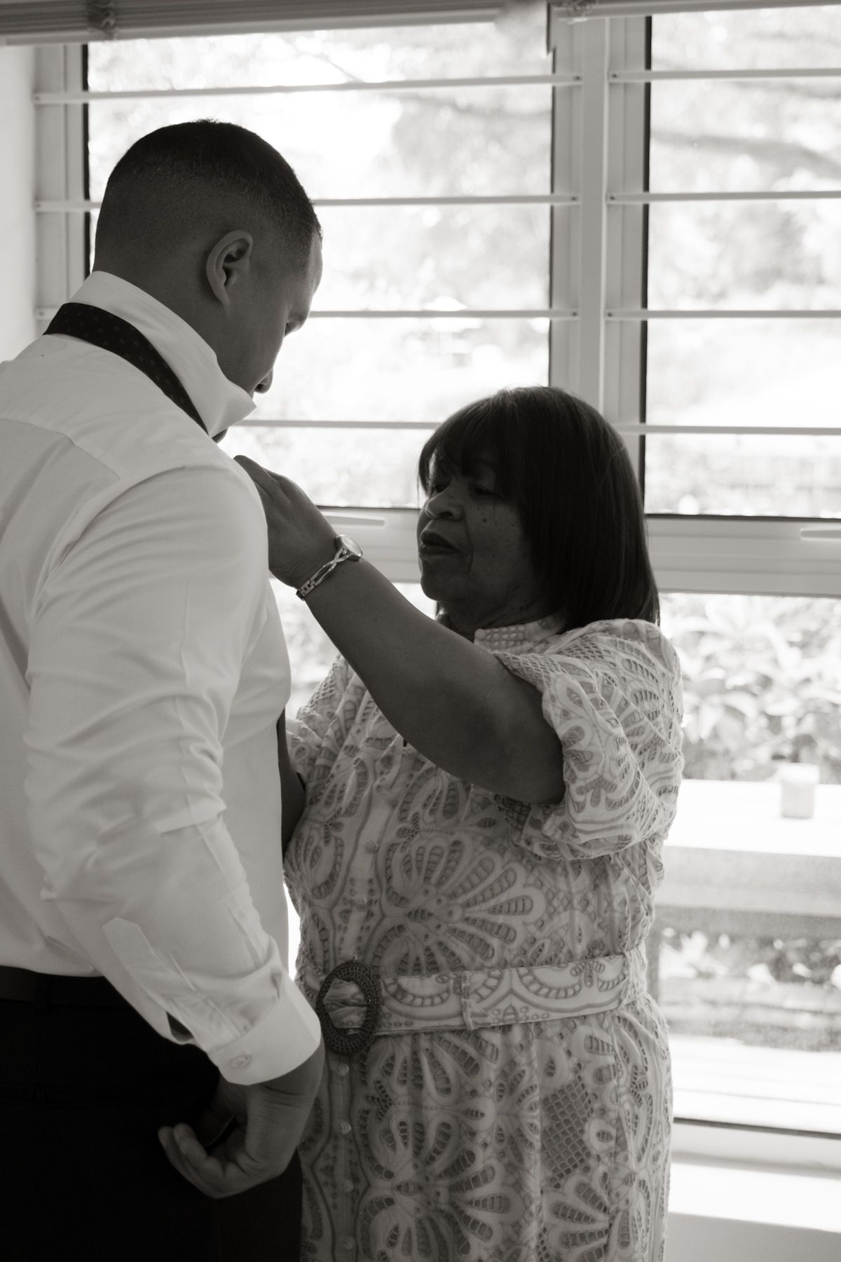 Black and white photo of mother helping groom with suit jacket before wedding ceremony.