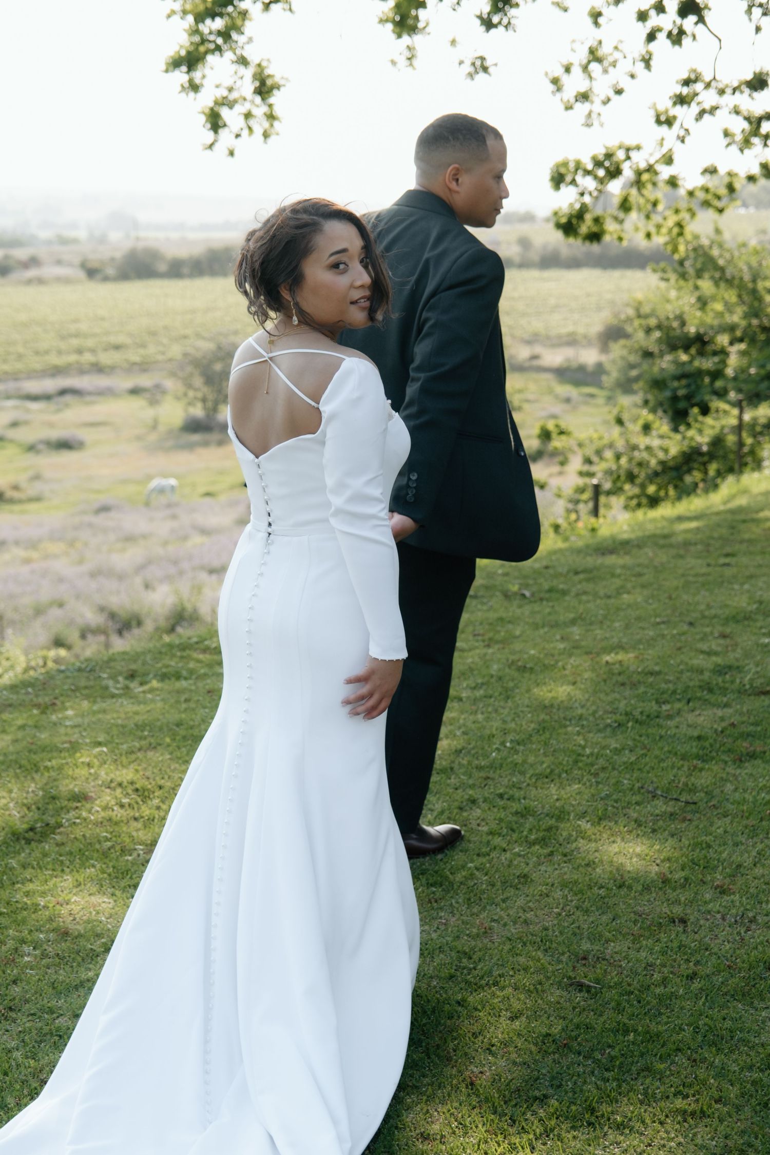 Bride in a fitted white gown with cross-back detail smiles over her shoulder while groom stands beside her outdoors.