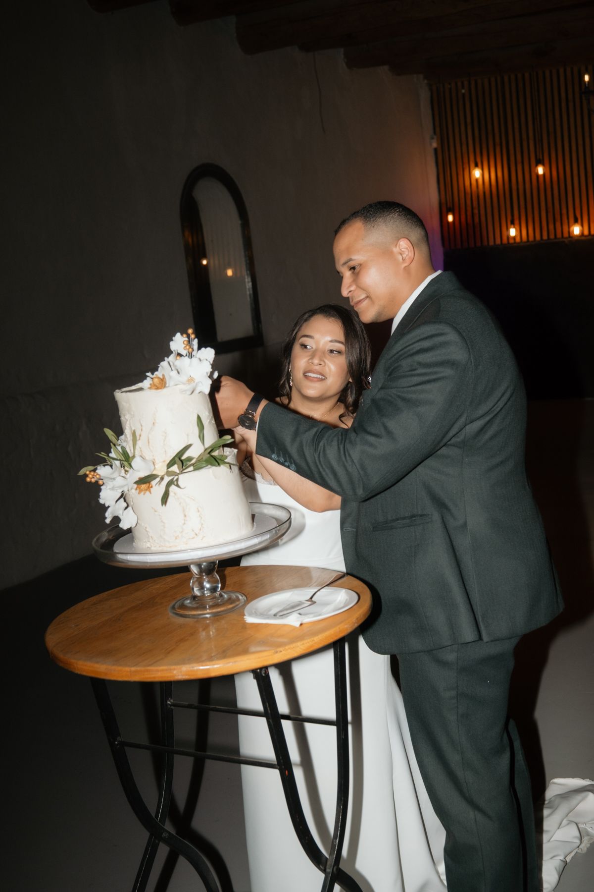 Smiling bride and groom cut elegant white floral wedding cake at their evening reception.