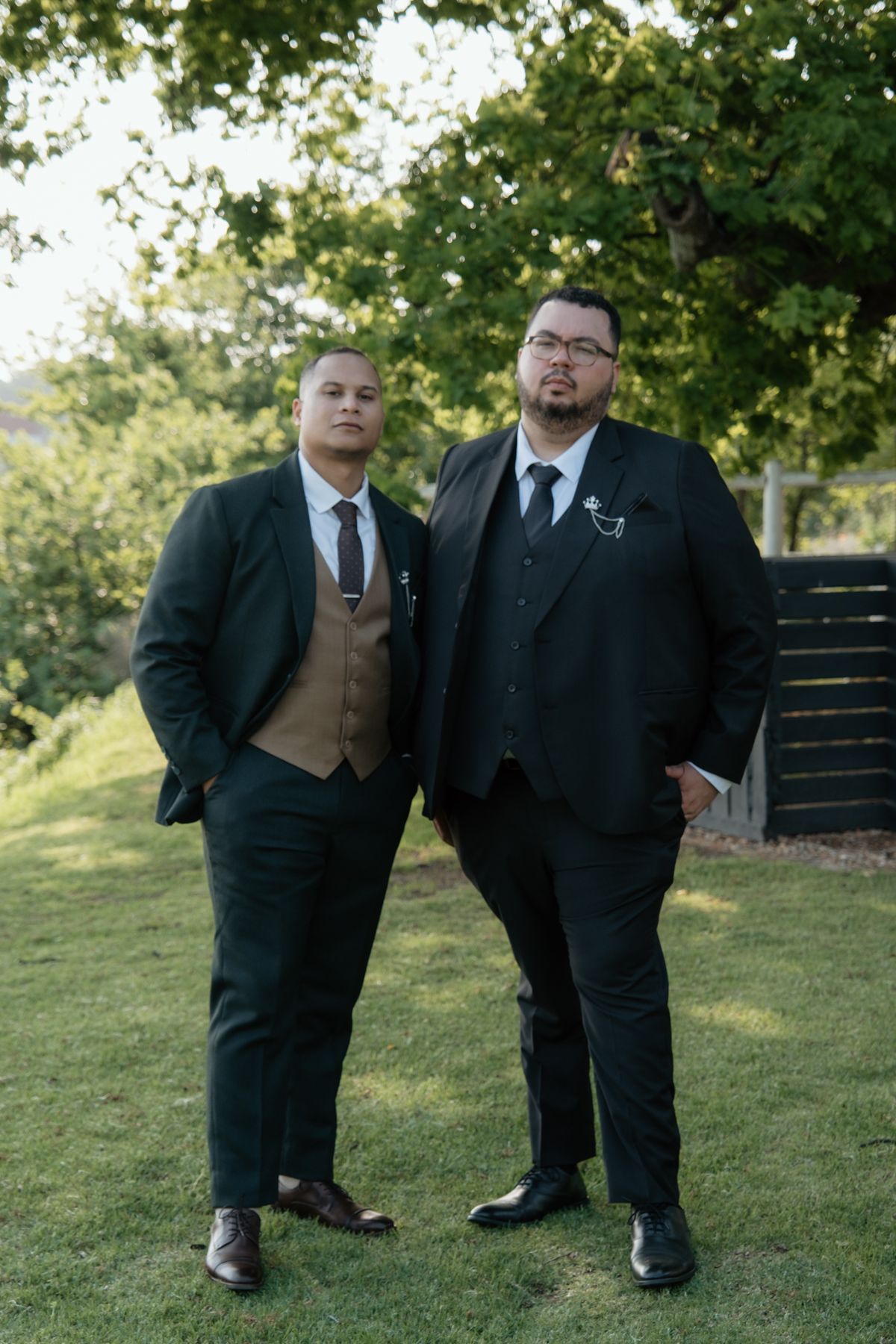 Two groomsmen in dark suits pose confidently on green lawn beneath large shade tree.