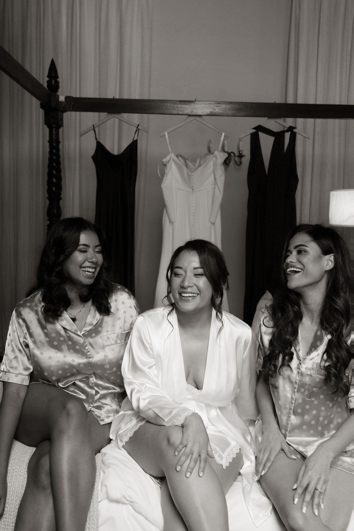 Three women in satin robes laugh together on a bed with wedding dresses hanging in the background.