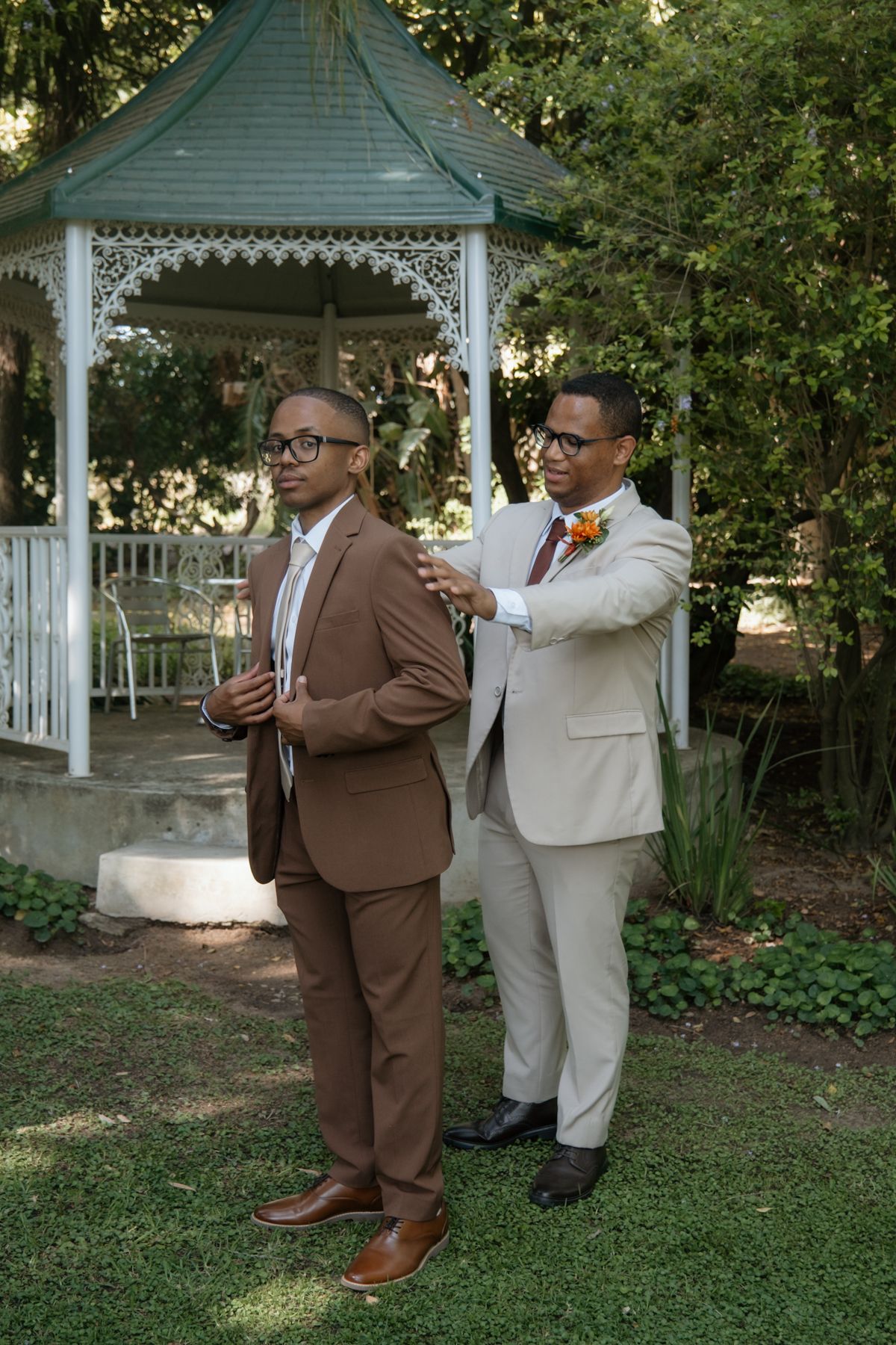 Groom in brown suit gets jacket adjusted by groomsman in tan suit near white gazebo outdoors.