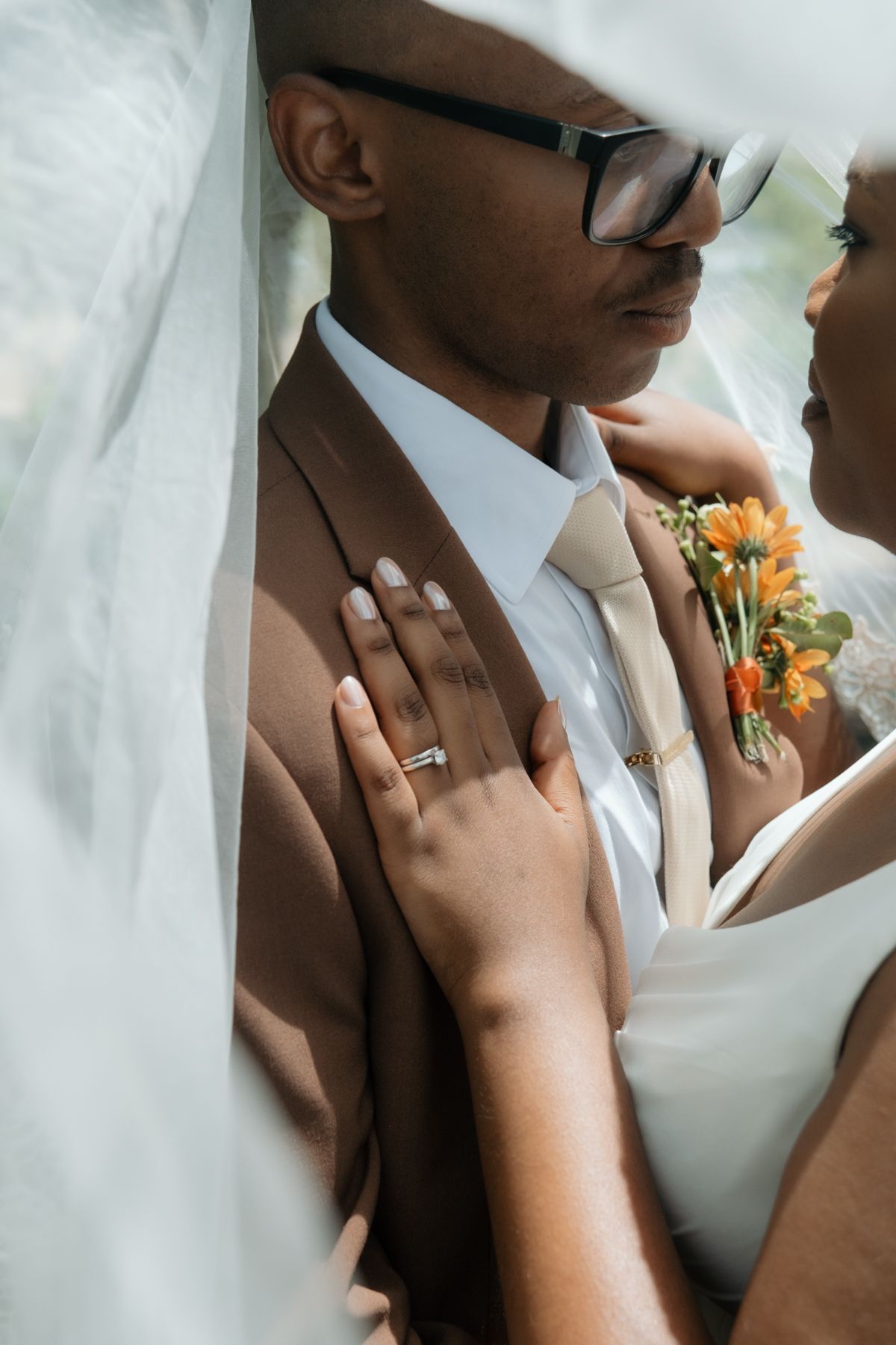 A bride and groom share an intimate moment, she wears a diamond ring and holds orange flowers against his brown suit.