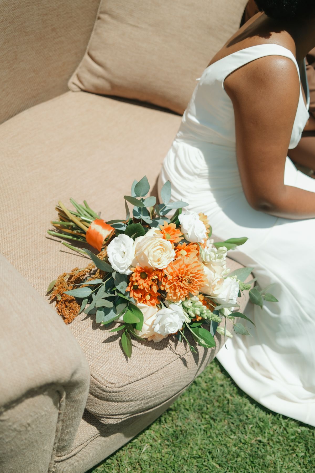 Bride in white gown sitting on beige sofa with orange and white wedding bouquet featuring dahlias and roses.
