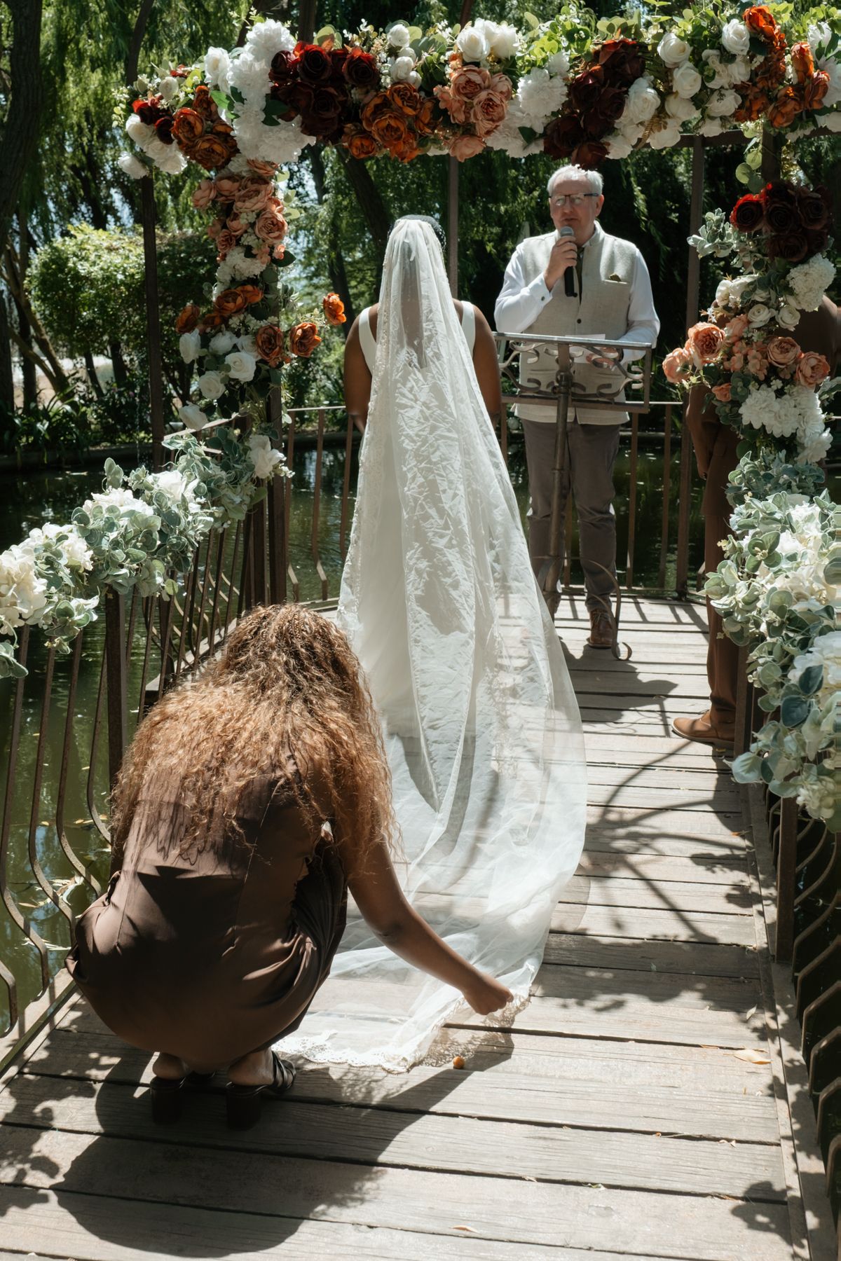 Wedding assistant adjusts bride's lace veil train on decorated floral bridge as officiant waits at altar.