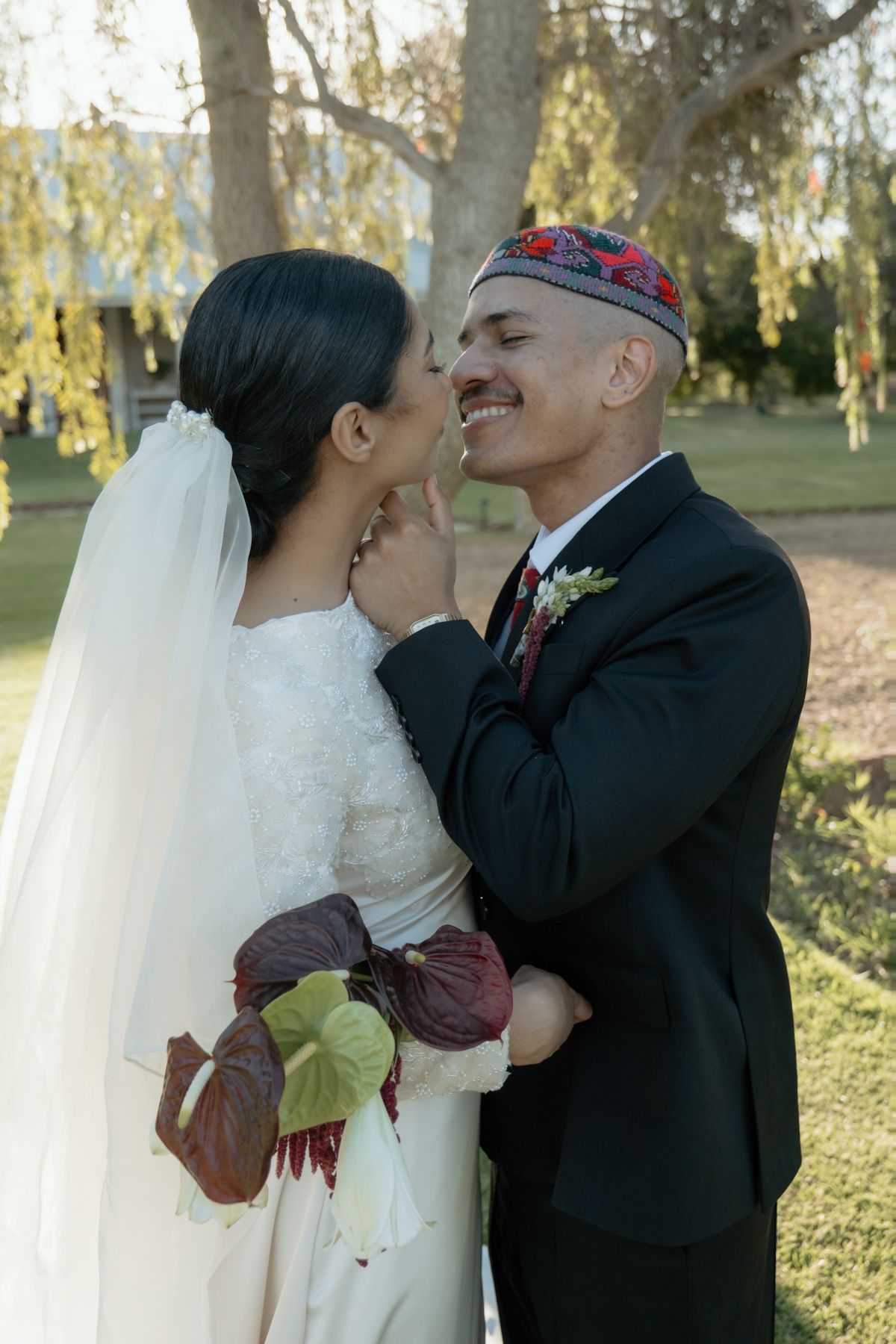 Smiling bride and groom in intimate moment outdoors, groom wearing embroidered kufi cap and boutonniere.