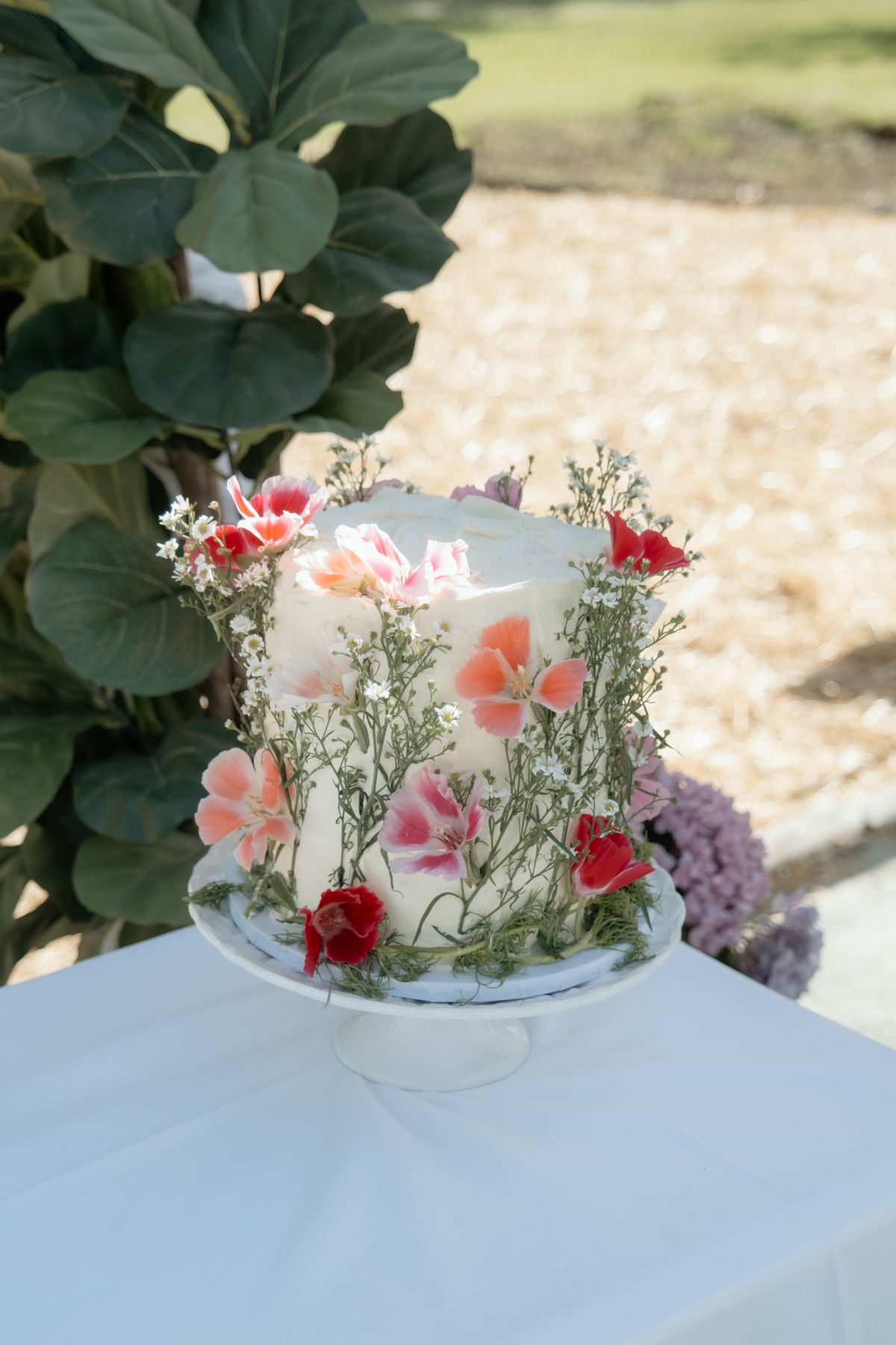 Elegant one-tier white wedding cake decorated with colorful pressed flowers and greenery on a stand.