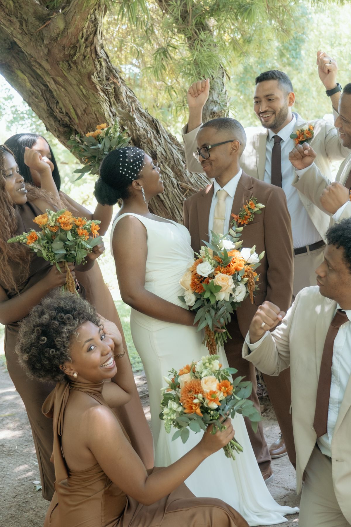 Smiling bride in white gown holds orange bouquet as wedding party cheers around her under a shady tree.