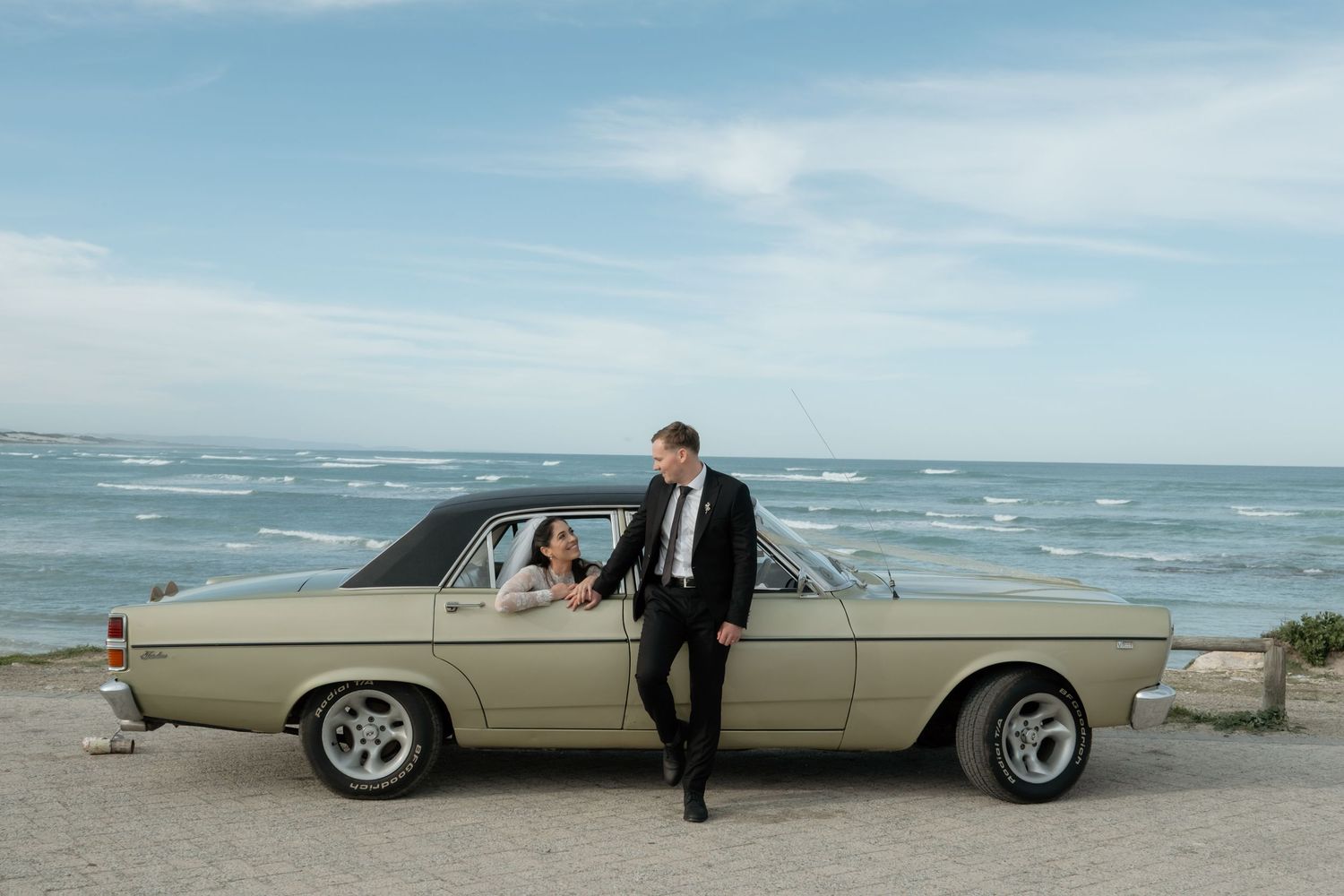 Bride and groom posing beside a vintage cream car by the ocean on their wedding day.