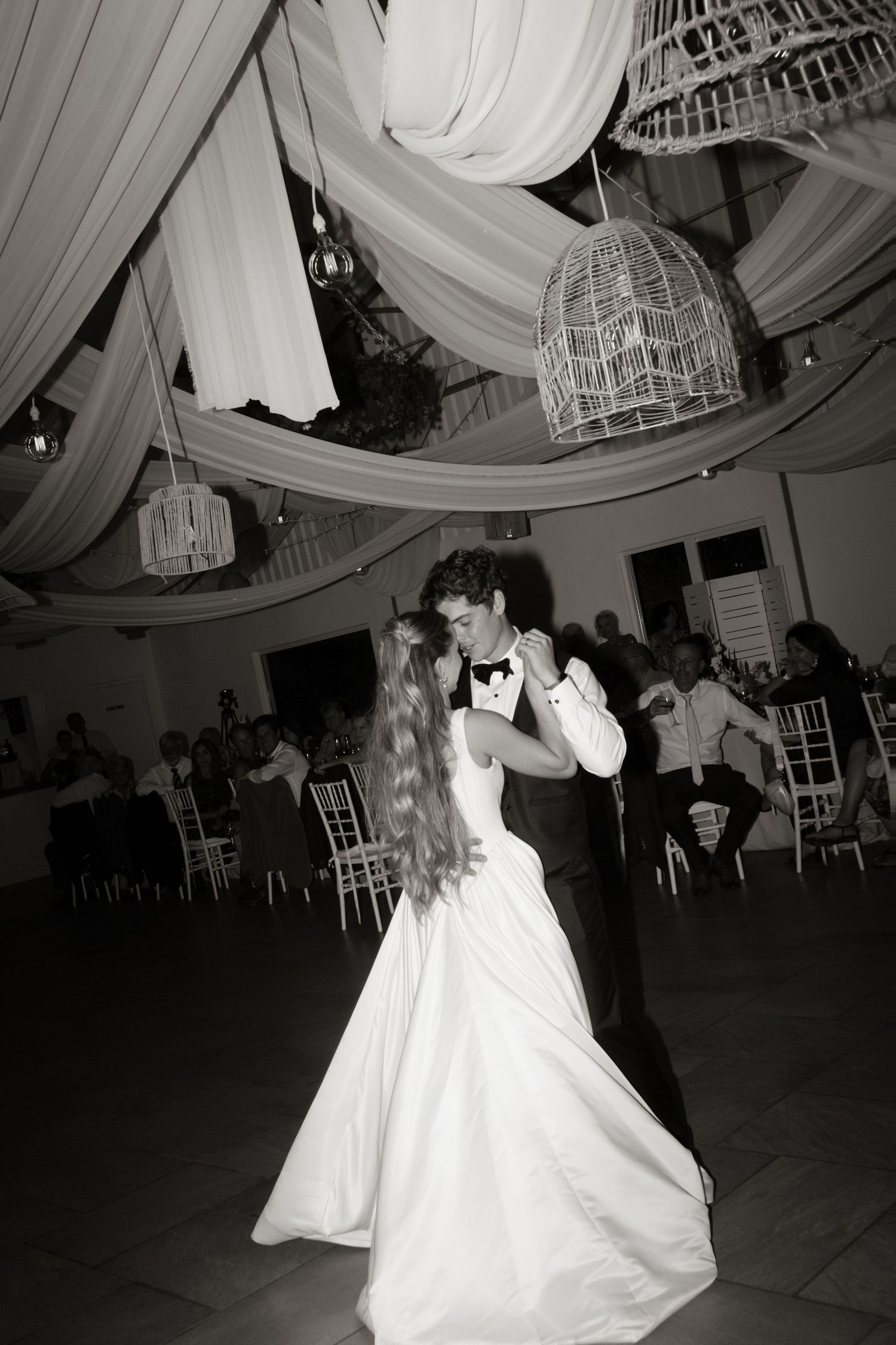 A black and white photo captures a couple dancing beneath draped fabric and birdcage chandeliers at a wedding reception.