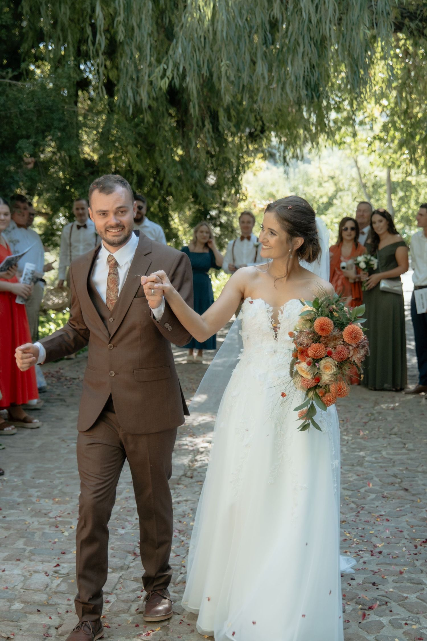 Smiling bride and groom holding hands walking through garden aisle lined with cheering wedding guests.