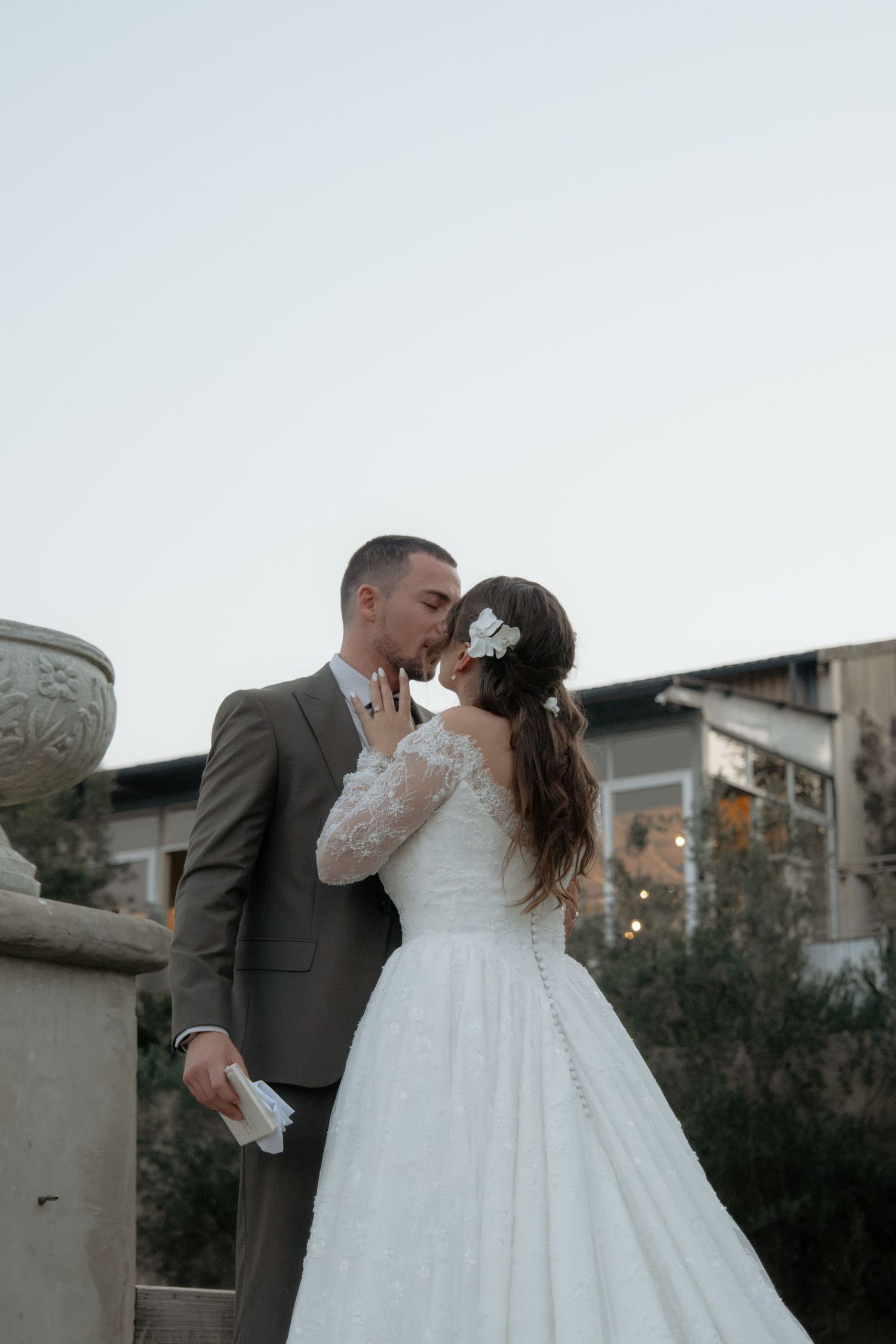 Bride and groom sharing a romantic kiss outdoors at dusk with decorative stone urns nearby.
