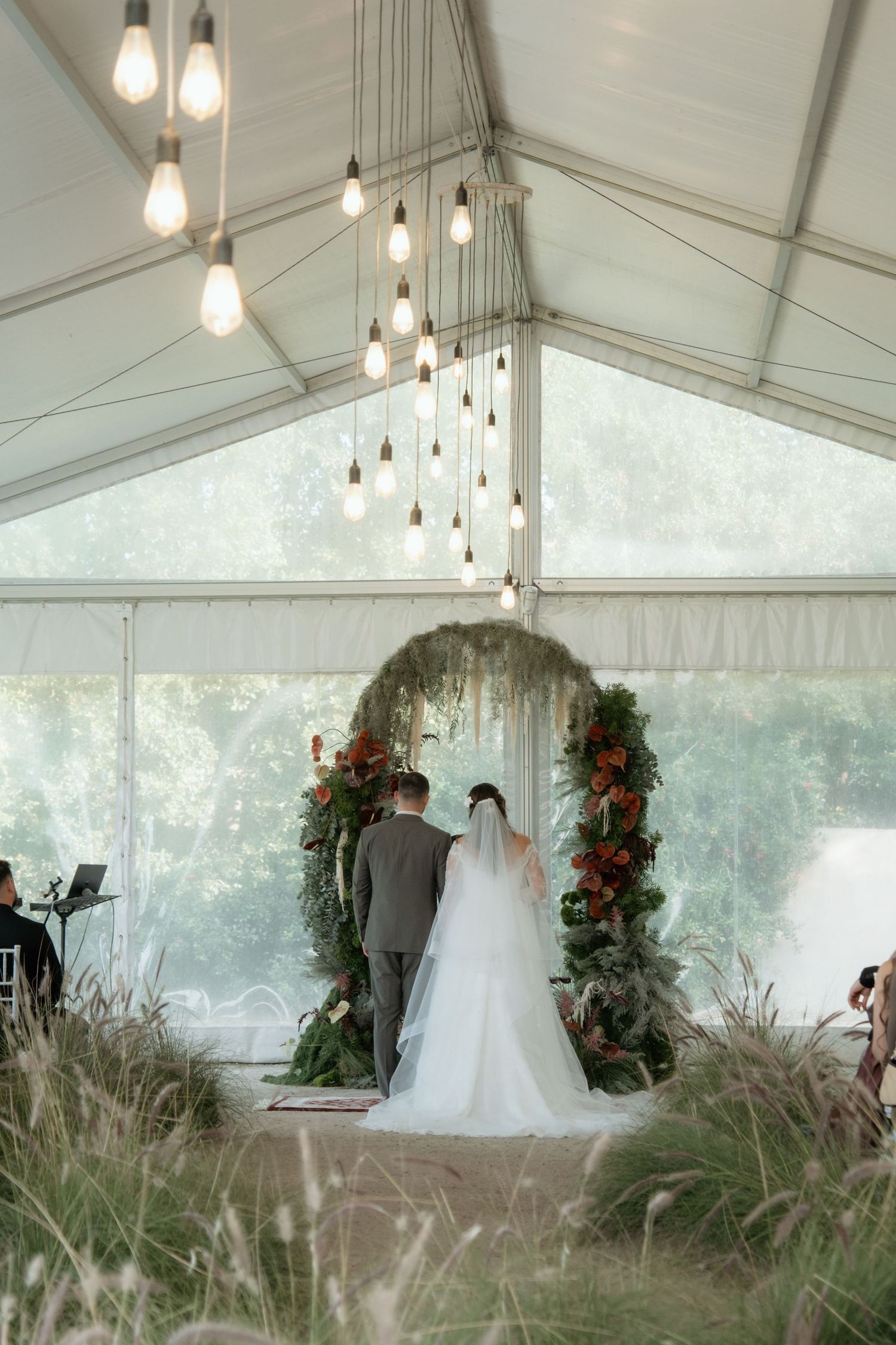 Bride and groom standing at circular floral arch under tent with hanging Edison bulbs.