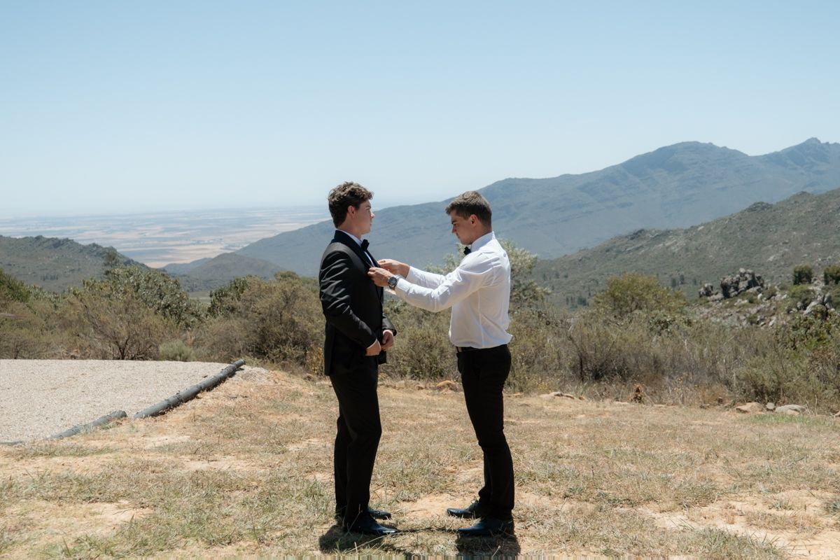 Two men in formal attire share a moment on a scenic mountain overlook under a clear blue sky.