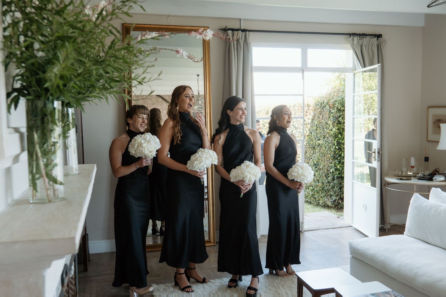 Four bridesmaids in matching black halter dresses holding white bouquets standing in bright modern room.
