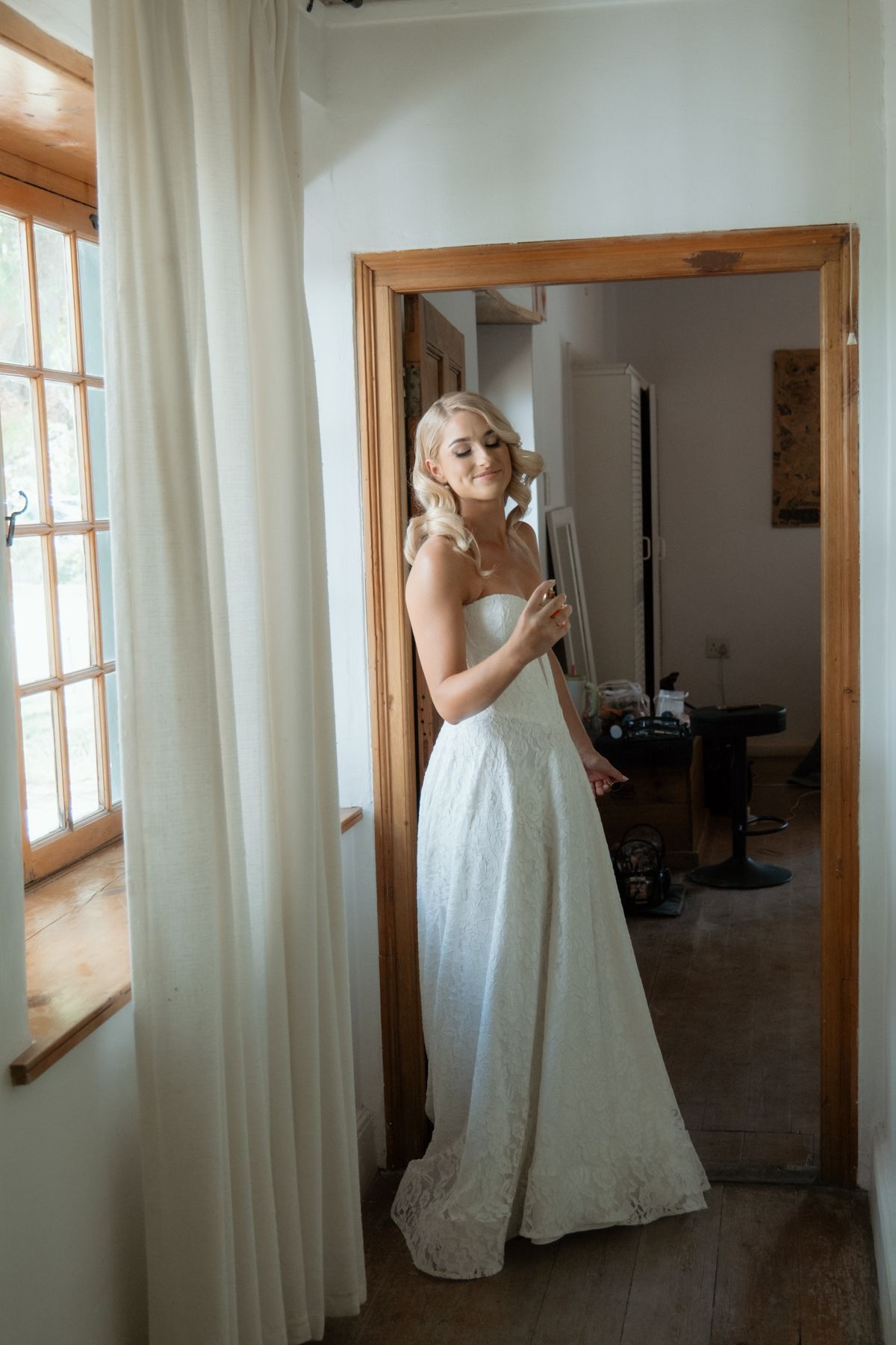 Bride in strapless white lace wedding gown standing in doorway of rustic venue getting ready.
