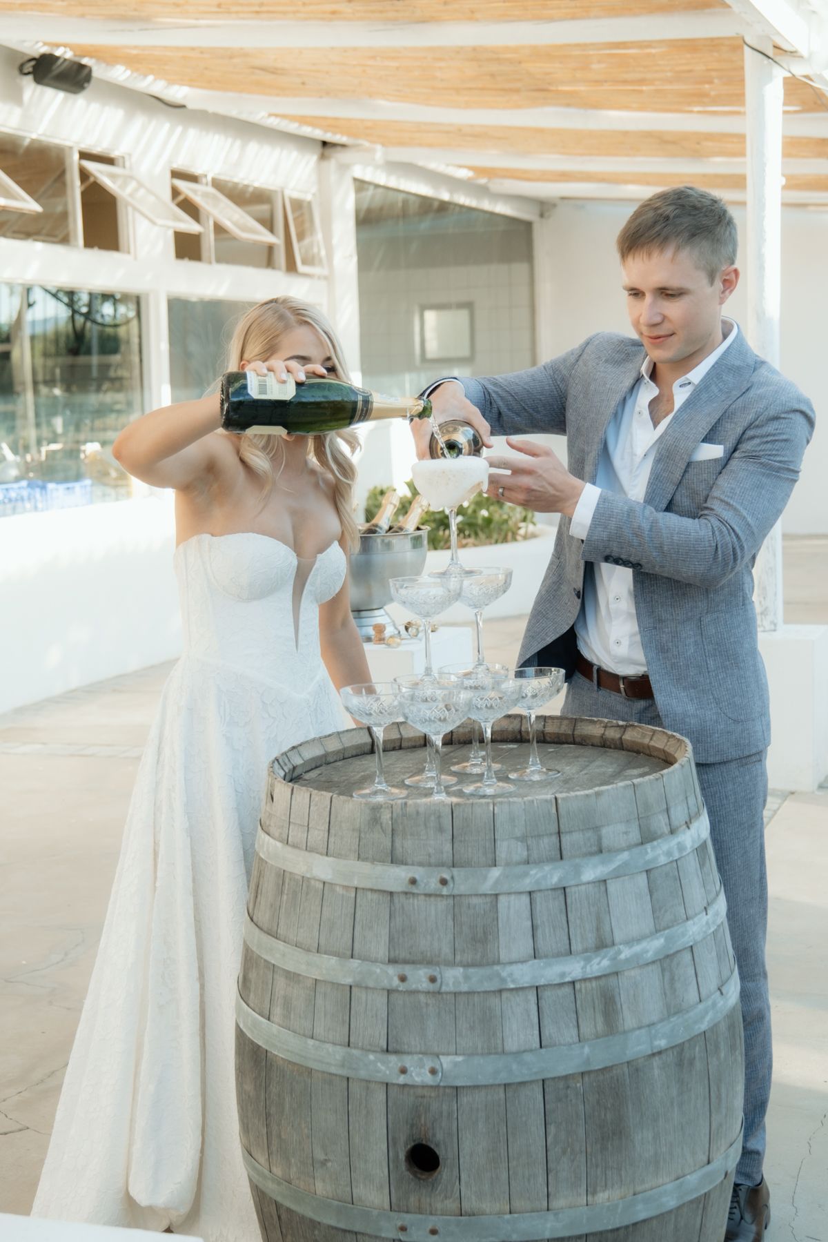 Bride and groom pour champagne tower on rustic wooden barrel at elegant outdoor wedding venue.