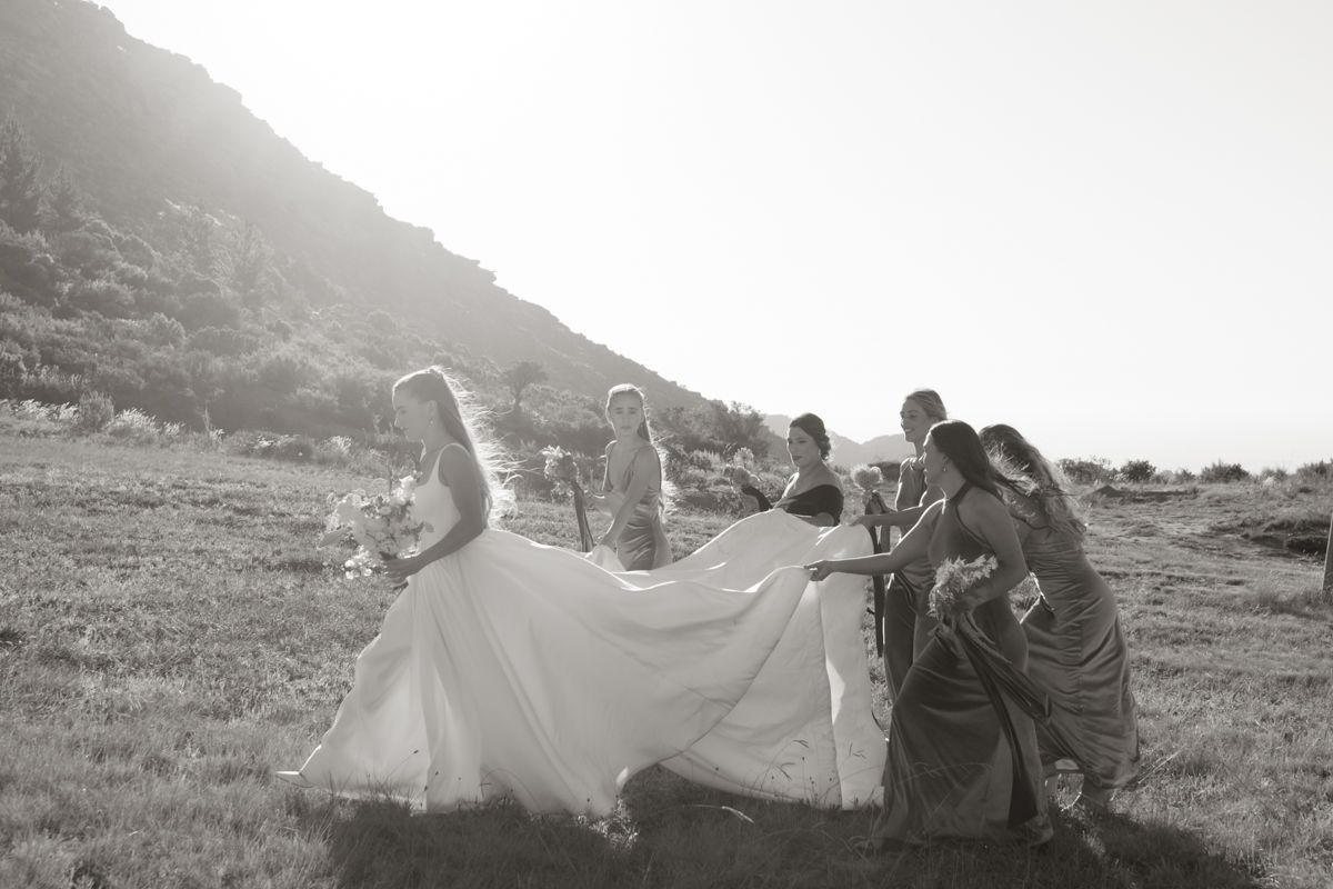 Black and white photo of bridesmaids helping bride with wedding gown train in scenic outdoor hillside setting.