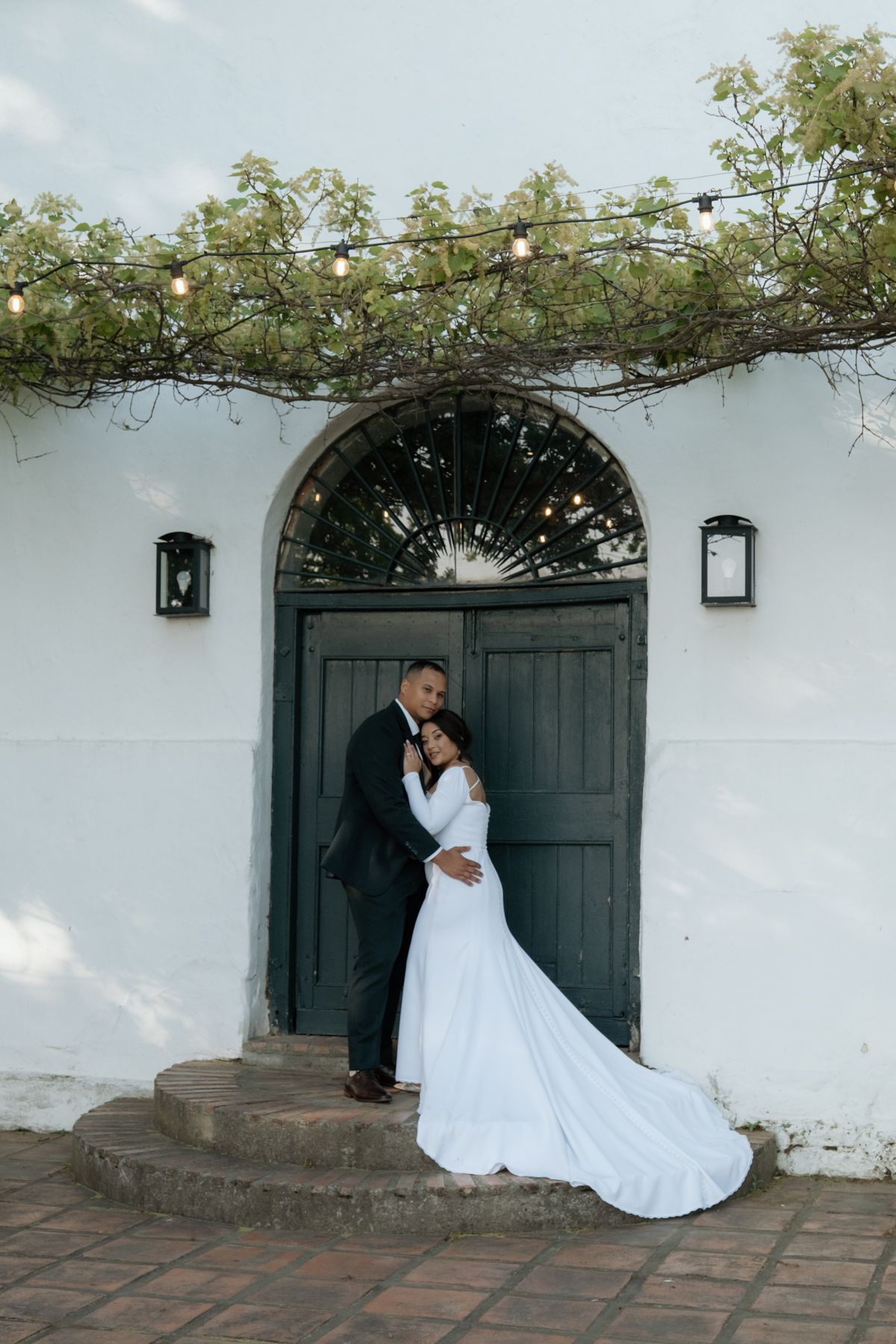 Bride and groom embracing before rustic white arched doorway with string lights at wedding venue.