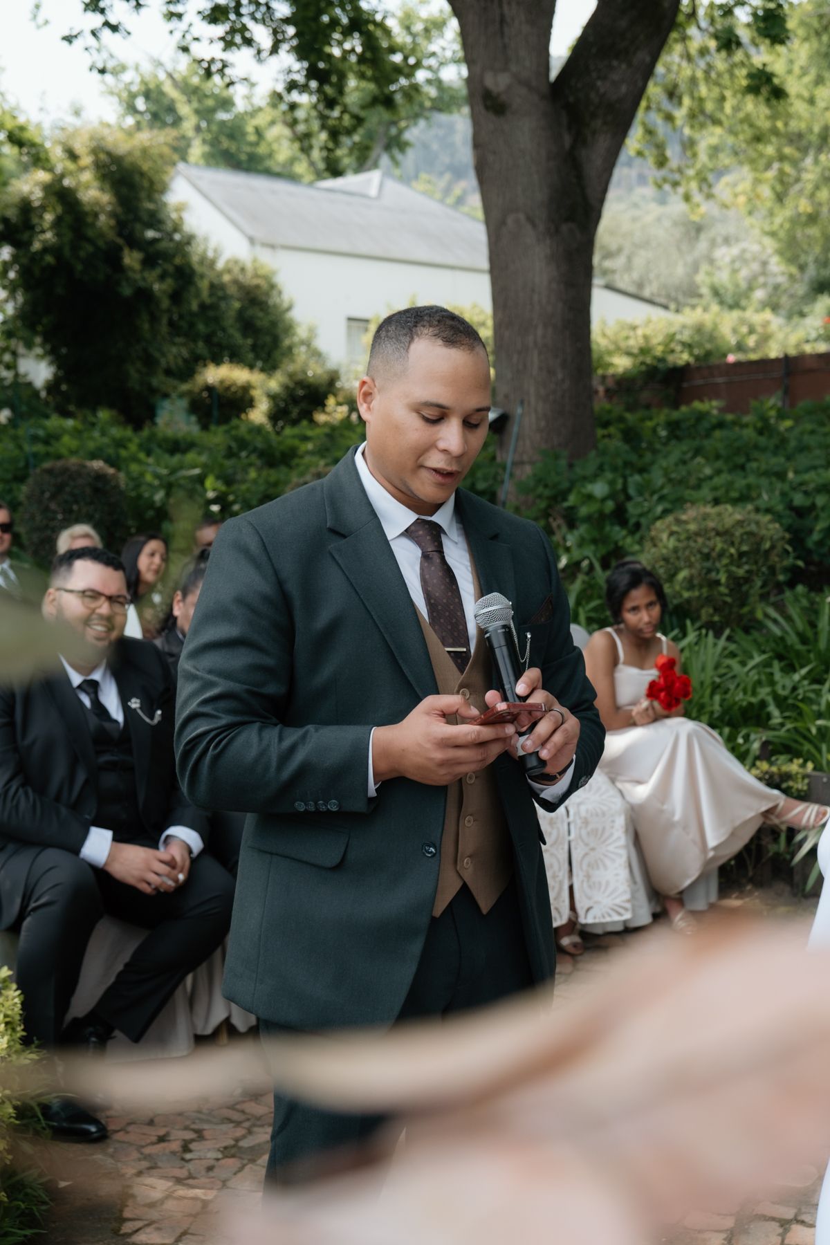 Groom in dark suit reading vows from phone during outdoor garden wedding ceremony.