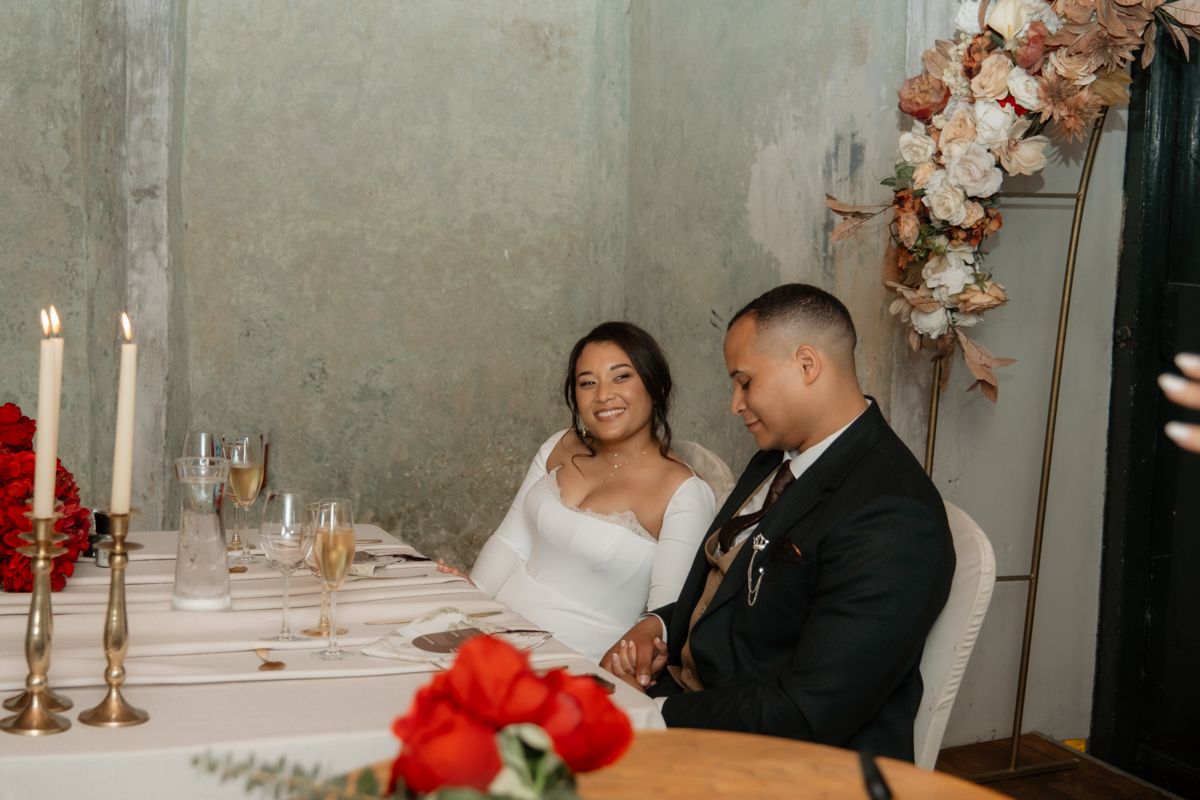 Bride and groom smile together at sweetheart table adorned with red roses, candles, and blush floral arrangements.