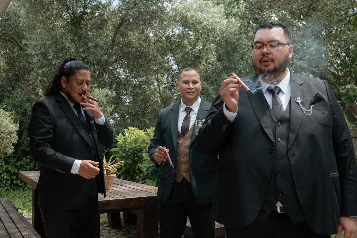 Three groomsmen in dark suits smoking cigars outdoors among olive trees before the wedding.