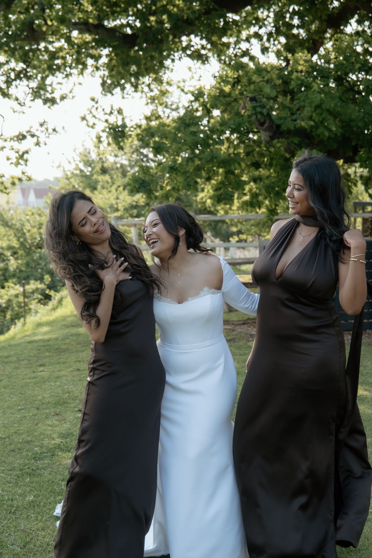 Bride in white gown laughing with two bridesmaids in dark brown satin dresses under a green tree.