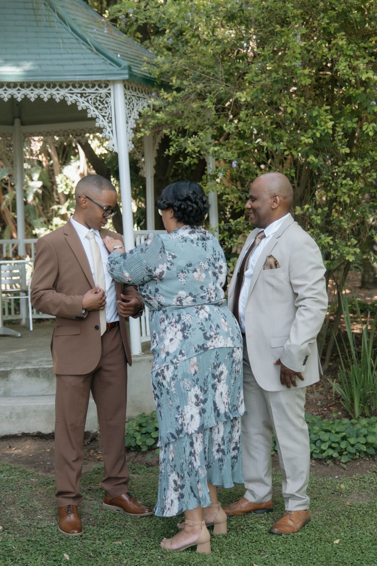 Three elegantly dressed wedding guests conversing near a white gazebo in a lush garden setting.