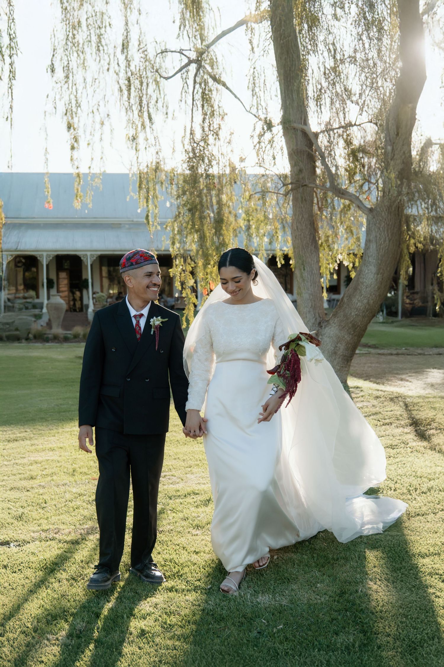 Bride and groom walk hand-in-hand on green lawn beneath willow tree, she in white gown, he in black suit with red cap.