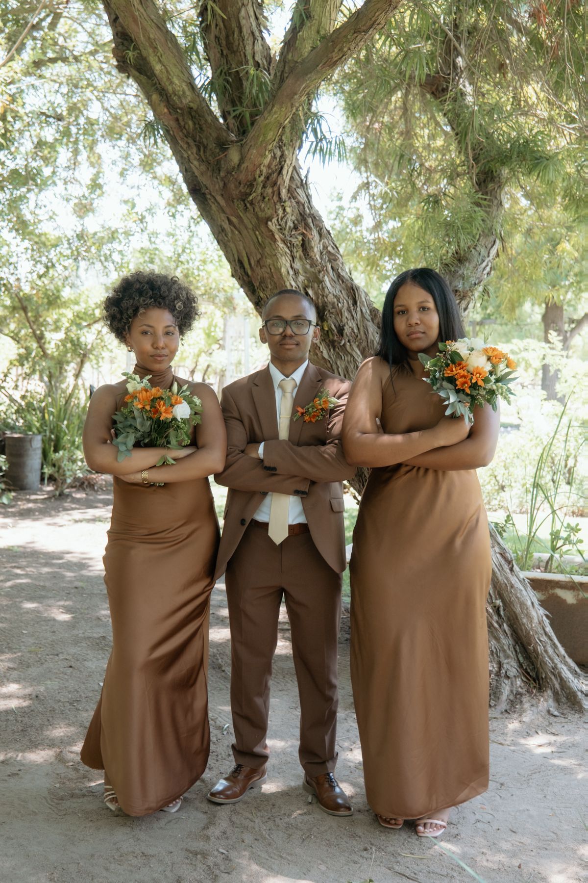 Groom in brown suit poses seriously with two bridesmaids in brown gowns holding orange bouquets under a tree.