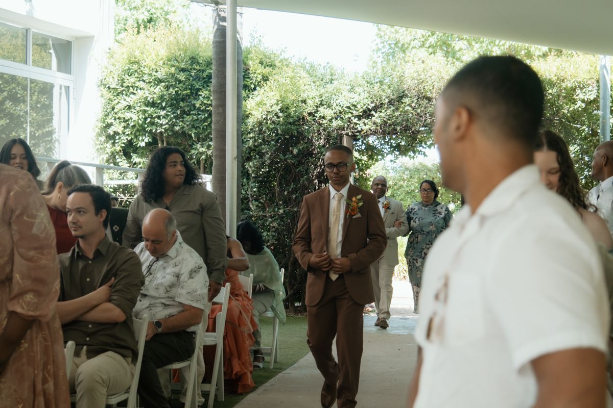 Guests gather at an outdoor wedding ceremony, with a man in a brown suit walking down the aisle.