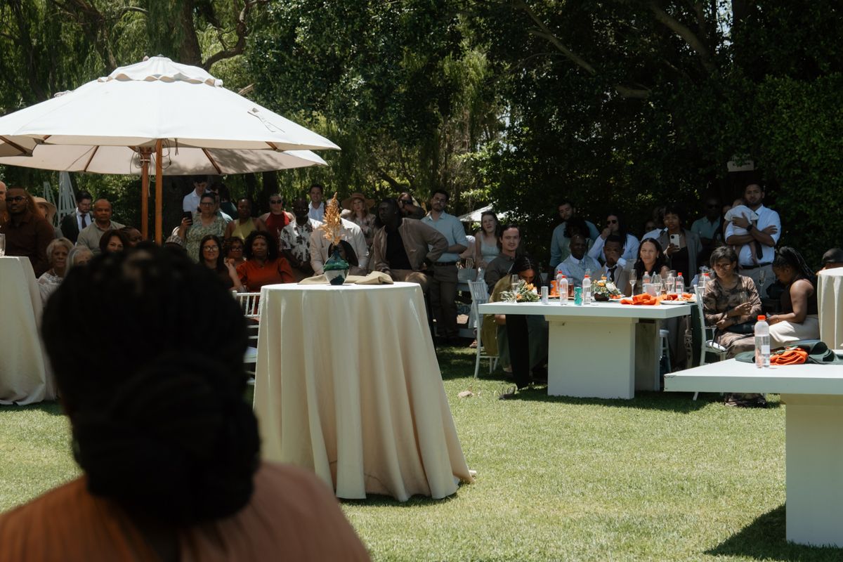 Outdoor garden gathering with guests seated at tables under a white umbrella on a sunny day.