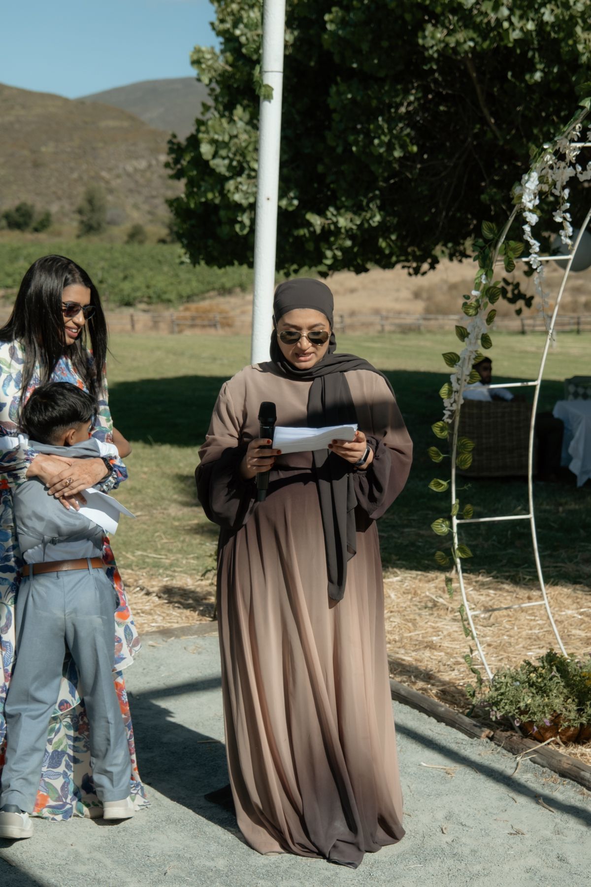 Woman in hijab reads speech at outdoor wedding ceremony with guests and scenic mountain backdrop.