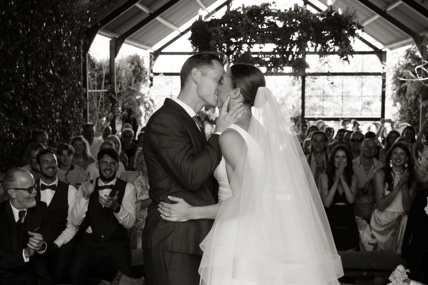 Black and white photo of a couple sharing their first kiss at a wedding ceremony surrounded by cheering guests.