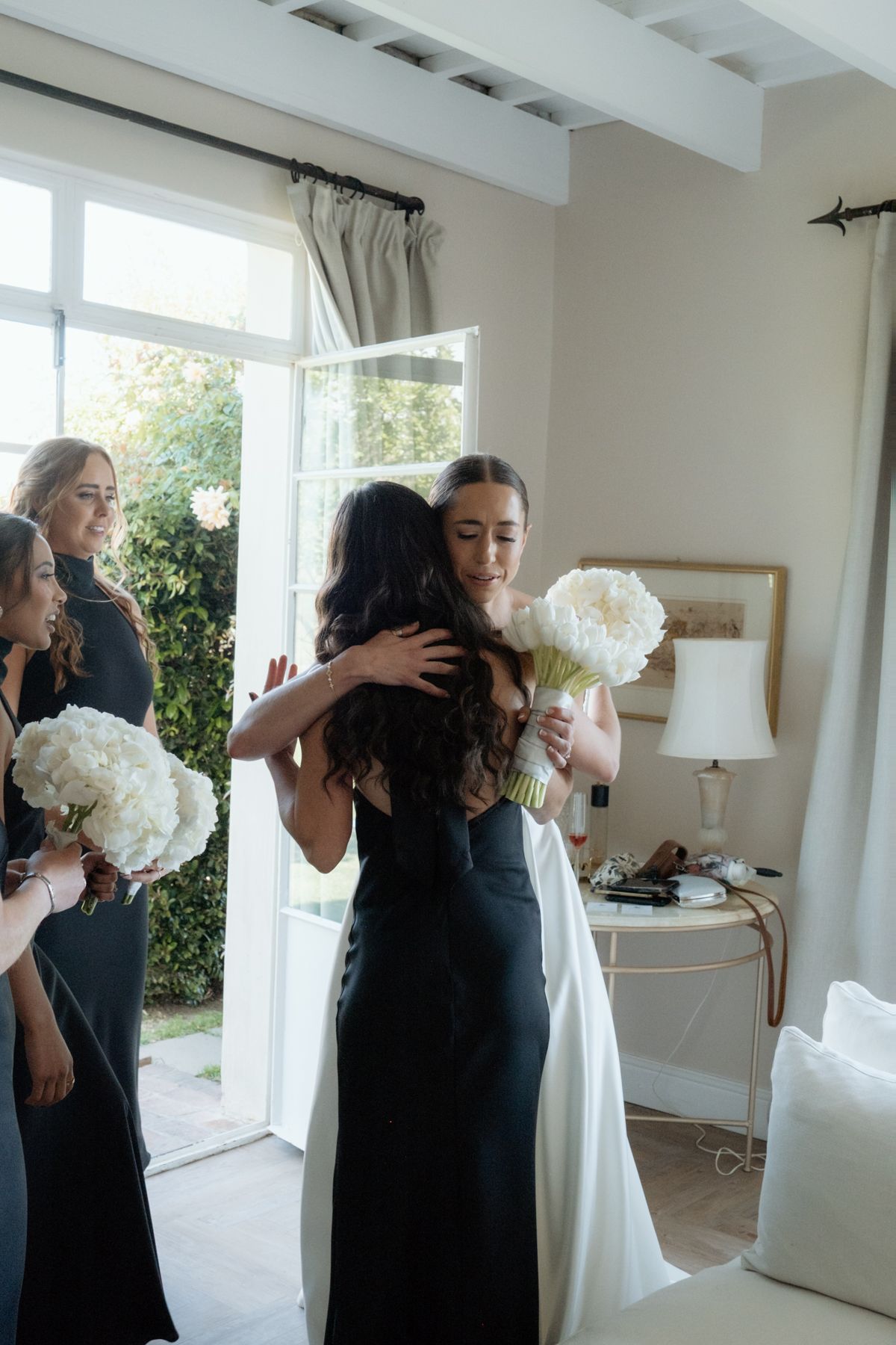 Bride in white dress embraces bridesmaid in black gown while others hold white peony bouquets indoors.