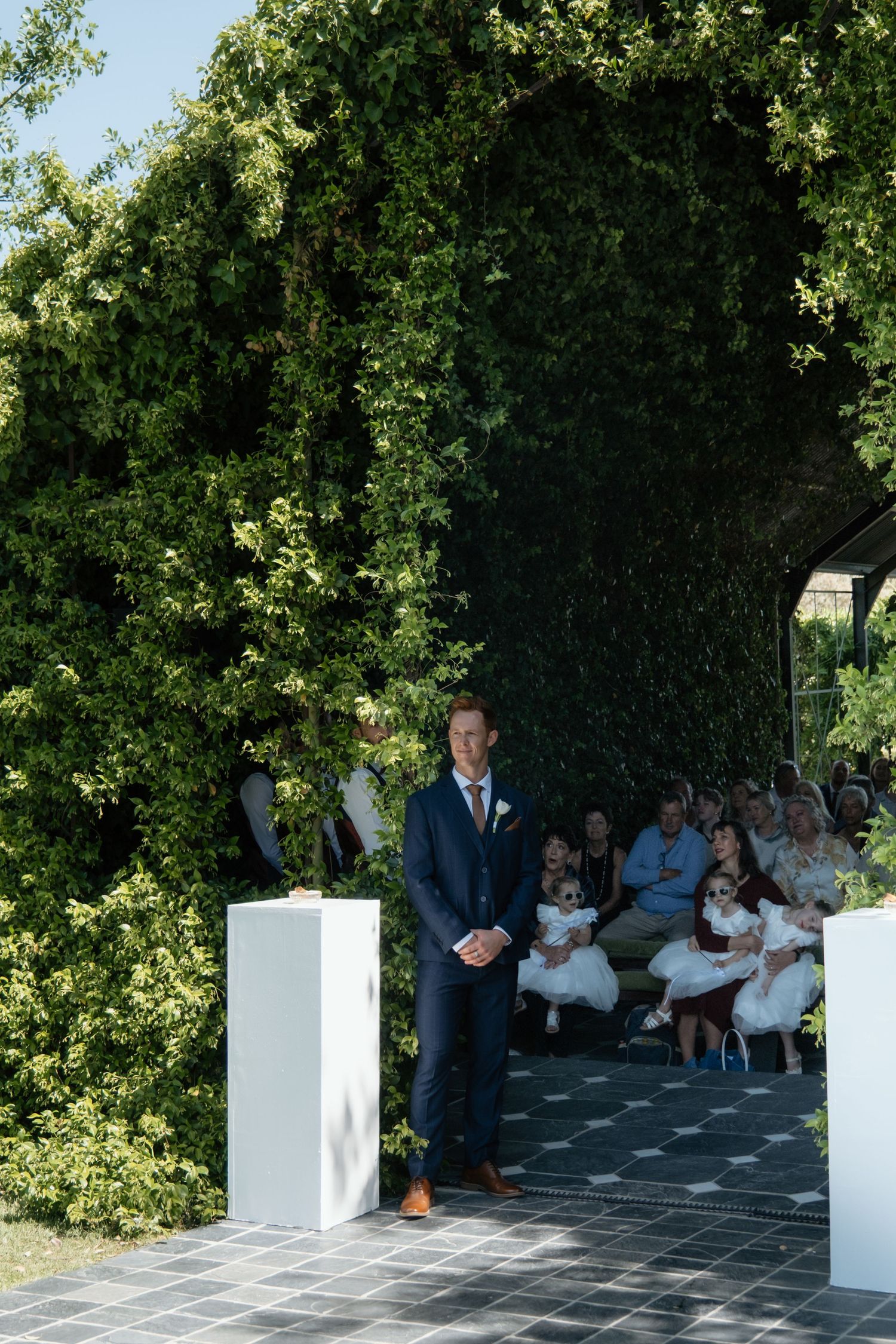 Groom in navy suit stands at floral arch altar awaiting bride at outdoor wedding ceremony.