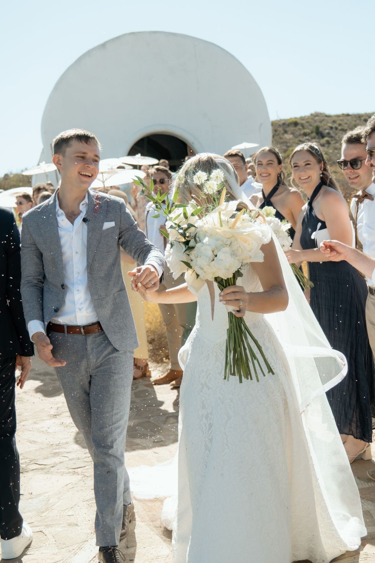 Groom in grey suit holds bride's hand as they exit a whitewashed chapel while guests toss confetti.