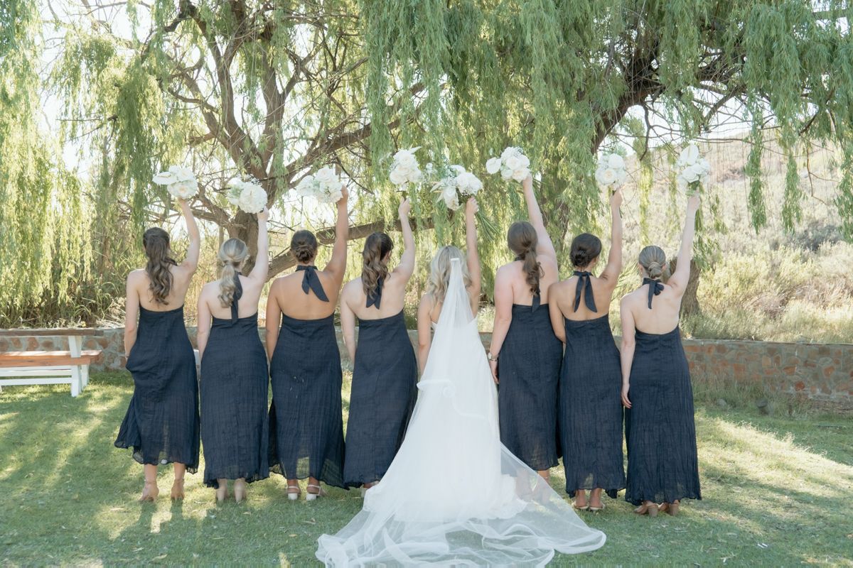 Bride and bridesmaids in navy dresses raising bouquets under a willow tree, viewed from behind.