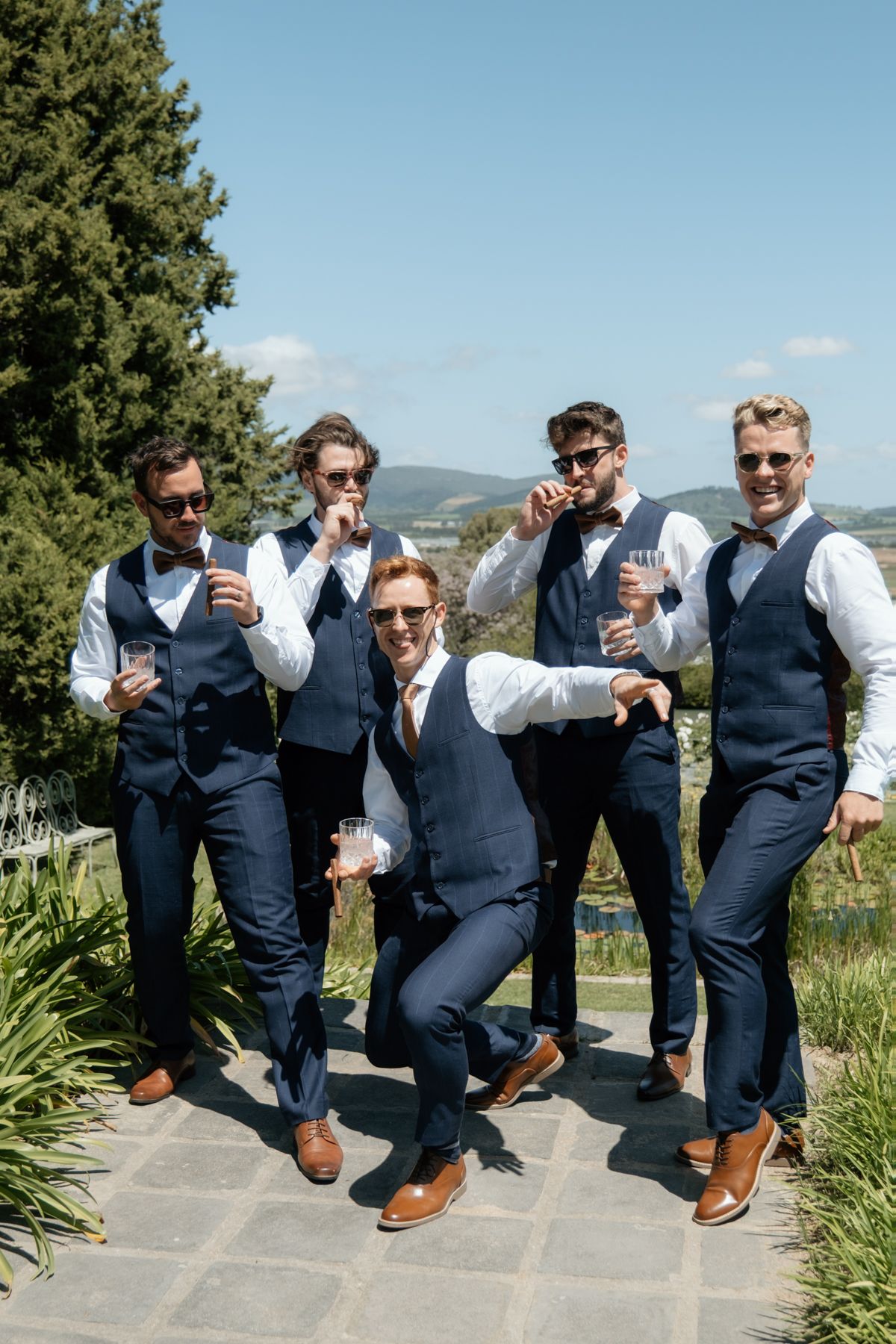 Five groomsmen in navy vests and bow ties pose playfully outdoors in sunny garden setting.